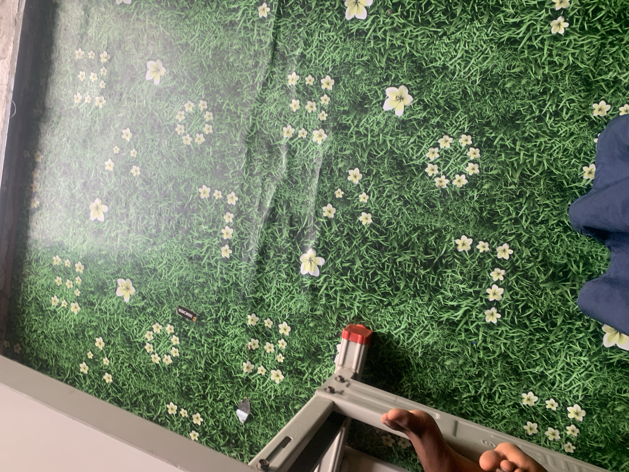 The image shows a close-up, high-angle view of a green, grassy wallpaper with scattered white and yellow flowers. A white ladder is positioned on the left side of the frame, with a person's bare foot resting on one of its steps. The person is wearing a dark blue garment. A black object with yellow lettering, possibly a battery, is visible on the grass. The wallpaper appears to be a backdrop or mural. The overall impression is of an indoor setting with a nature-themed decoration. The provided location, Jauro Yinu, Nigeria, is not evident from the image itself.