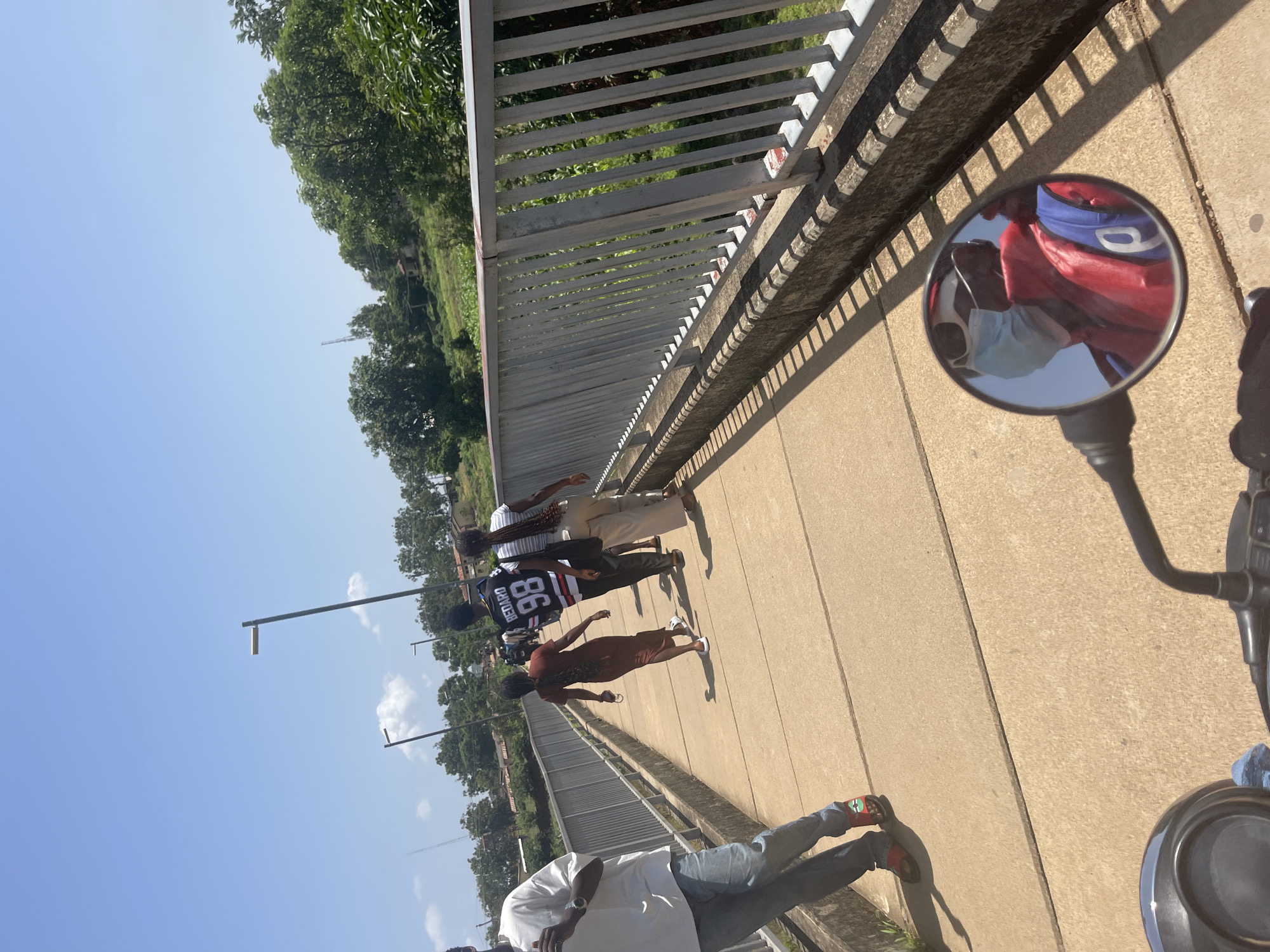 The image shows a concrete pedestrian bridge with a metal railing. Several people are walking on the bridge. On the left, a person in a white t-shirt and blue jeans is walking away from the camera. Further down the bridge, a person in a brown dress is walking away. Behind them, a person in a black and white jersey with the number "98" is walking, followed by a person in a striped shirt and light-colored pants. A motorcycle handlebar and mirror are visible in the foreground, with a reflection of a person wearing a light blue face mask and a red and blue jacket. The environment appears to be a sunny day with trees and buildings visible on the hillside in the background. The location is identified as Rafin Par, Nigeria.