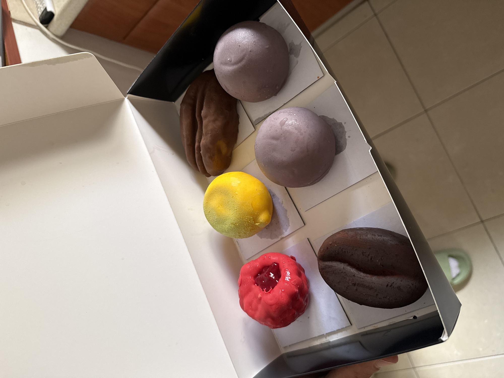 An elevated view captures an open cardboard box containing six distinct desserts. A hand is visible holding the box from the bottom right. The box features a white interior with individual compartments, and black exterior accents. Inside, arranged in two columns, are the confectionery items. On the left column, from top to bottom, are: a brown, elongated dessert with a ridged texture, resembling a large date or nut; a bright yellow, lemon-shaped dessert with a slightly frosty appearance; and a vibrant red, ring-shaped dessert with a dark red, jelly-like filling in its center. On the right column, from top to bottom, are: two smooth, spherical, lavender-colored desserts, and a dark brown dessert molded into the shape of a large coffee bean. Slight condensation is visible on the surface of several desserts, suggesting they are chilled or frozen. The background consists of light brown tiled flooring with visible grout lines, a section of a wooden counter or cabinet, a white cable, and a light green slipper partially visible on the floor. The scene takes place indoors, likely in a home in Ramat Gan, Israel. No text is visible in the image.