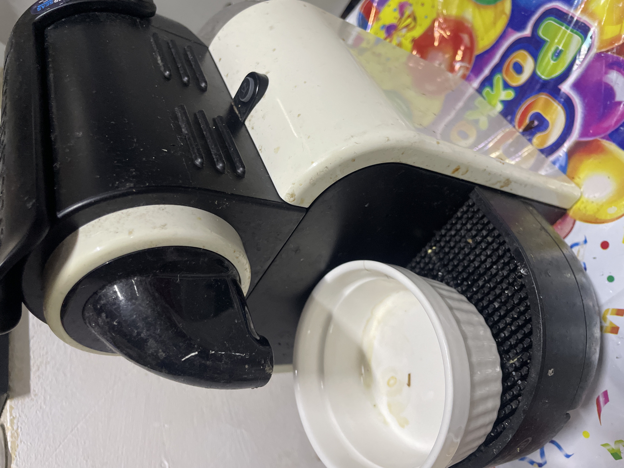 The image shows a close-up, angled view of a used coffee maker with a white and black design. A small white ramekin sits in the drip tray of the coffee maker. The coffee maker is positioned on a surface covered with colorful party decorations, including balloons and streamers, with visible text such as "OKTO" and partial letters from numbers. The background appears to be a wall. The overall scene suggests a post-party or informal setting. There are no people visible. The lighting is artificial and somewhat harsh, casting shadows. The coffee maker appears to be somewhat dirty, with visible residue. The location is Taganrog, Russia.