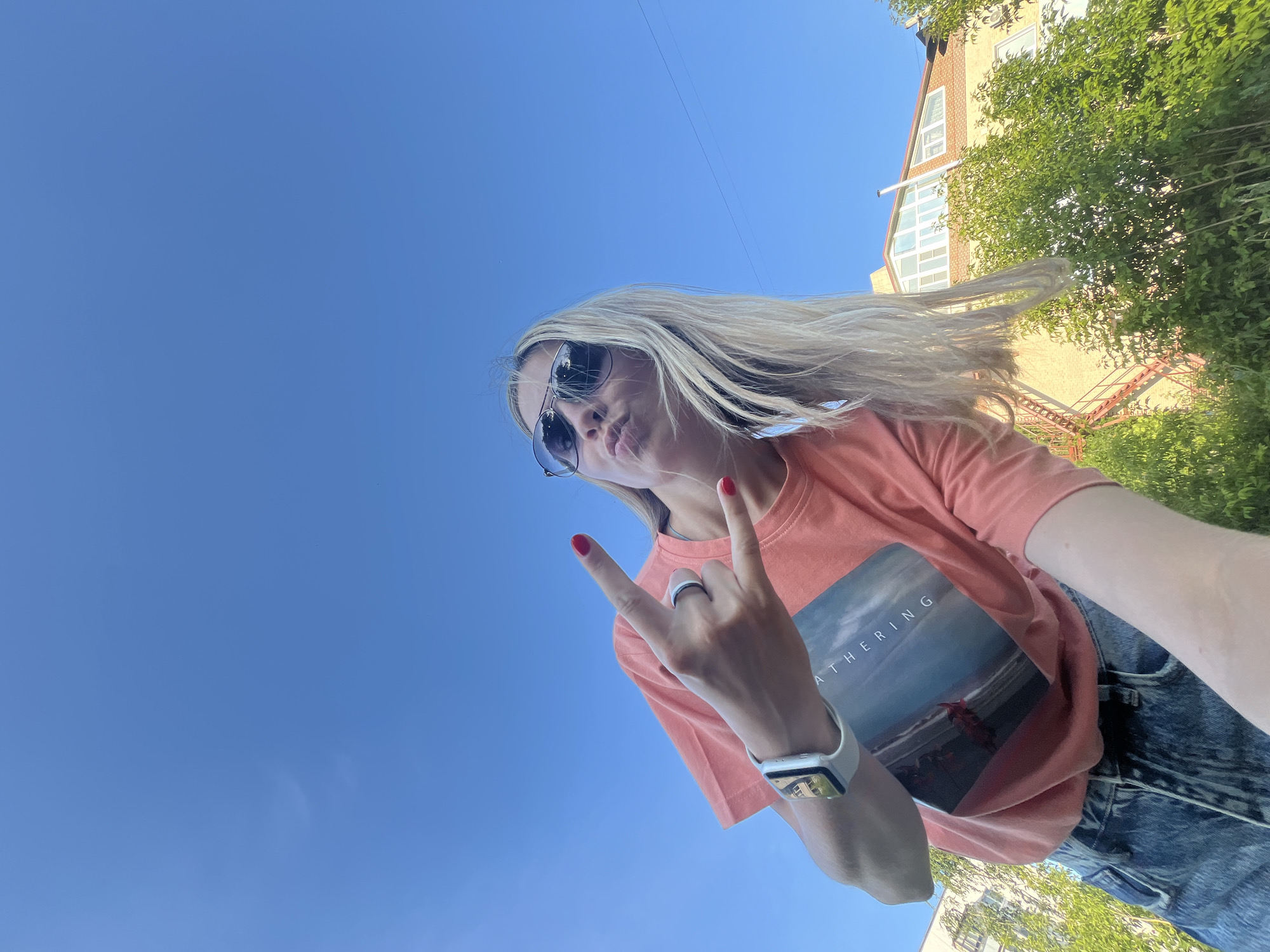 A low-angle, full-body outdoor selfie captures a young, fair-skinned woman with blonde hair, wearing sunglasses and a coral-colored t-shirt with a graphic print. She is making a "rock on" hand gesture with her right hand, displaying red nail polish. The woman is also wearing blue denim jeans and a white smartwatch on her left wrist. Her lips are pursed, as if blowing a kiss. The background is dominated by a clear, bright blue sky. In the lower right portion of the image, a building with a red-tiled roof and visible windows is partially obscured by lush green foliage. To the left, another building with a light-colored facade is visible. The scene appears to be set in a residential area of Chaikovsky, Russia, on a sunny day.
