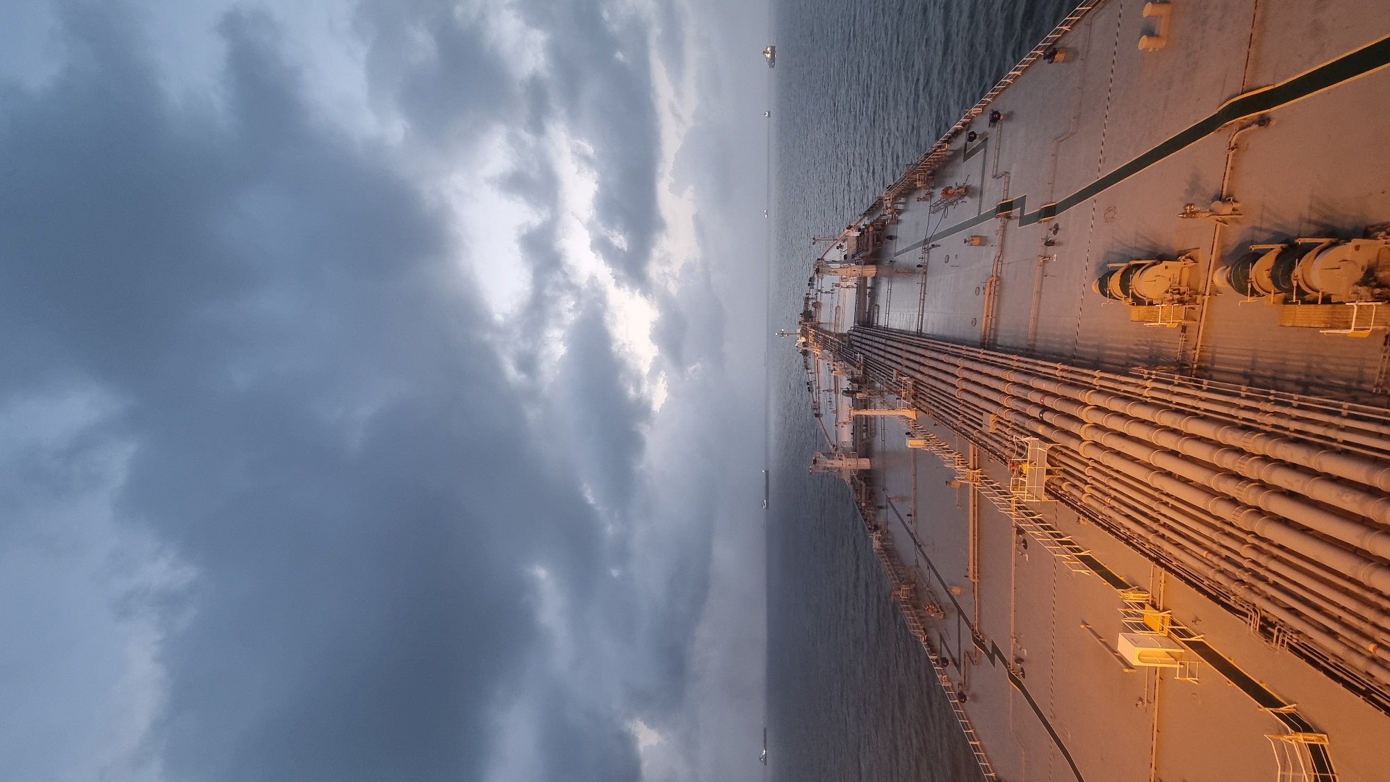 The image captures a view from the deck of a large vessel, likely a tanker, at sea. The sky is overcast with heavy, dark clouds dominating the upper portion of the frame, suggesting an approaching storm or twilight conditions. The sea is a deep, dark blue, with calm water indicating a lack of strong waves. Several other ships are visible as distant silhouettes on the horizon, scattered across the vast expanse of the ocean.
The foreground of the image shows the deck of the vessel, which is a light gray color. A prominent network of large, parallel pipes runs down the center of the deck, illuminated by a warm, golden light, possibly from the vessel's own lighting system. There are also various pieces of industrial equipment, such as winches and what appear to be pump housings, on the deck. Metal railings and gangways are also visible, demarcating pathways and sections of the deck.
The scene is devoid of any individuals actively engaged in visible actions. The overall impression is one of industrial maritime activity in a potentially challenging weather environment. The mention of Pirotan, India, in the prompt suggests the vessel might be operating in the waters surrounding this island, which is known for its marine national park and is part of the Gujarat Maritime Board. However, the image itself does not contain any specific visual cues to confirm this exact location, other than the possibility of the vessel being in Indian waters.