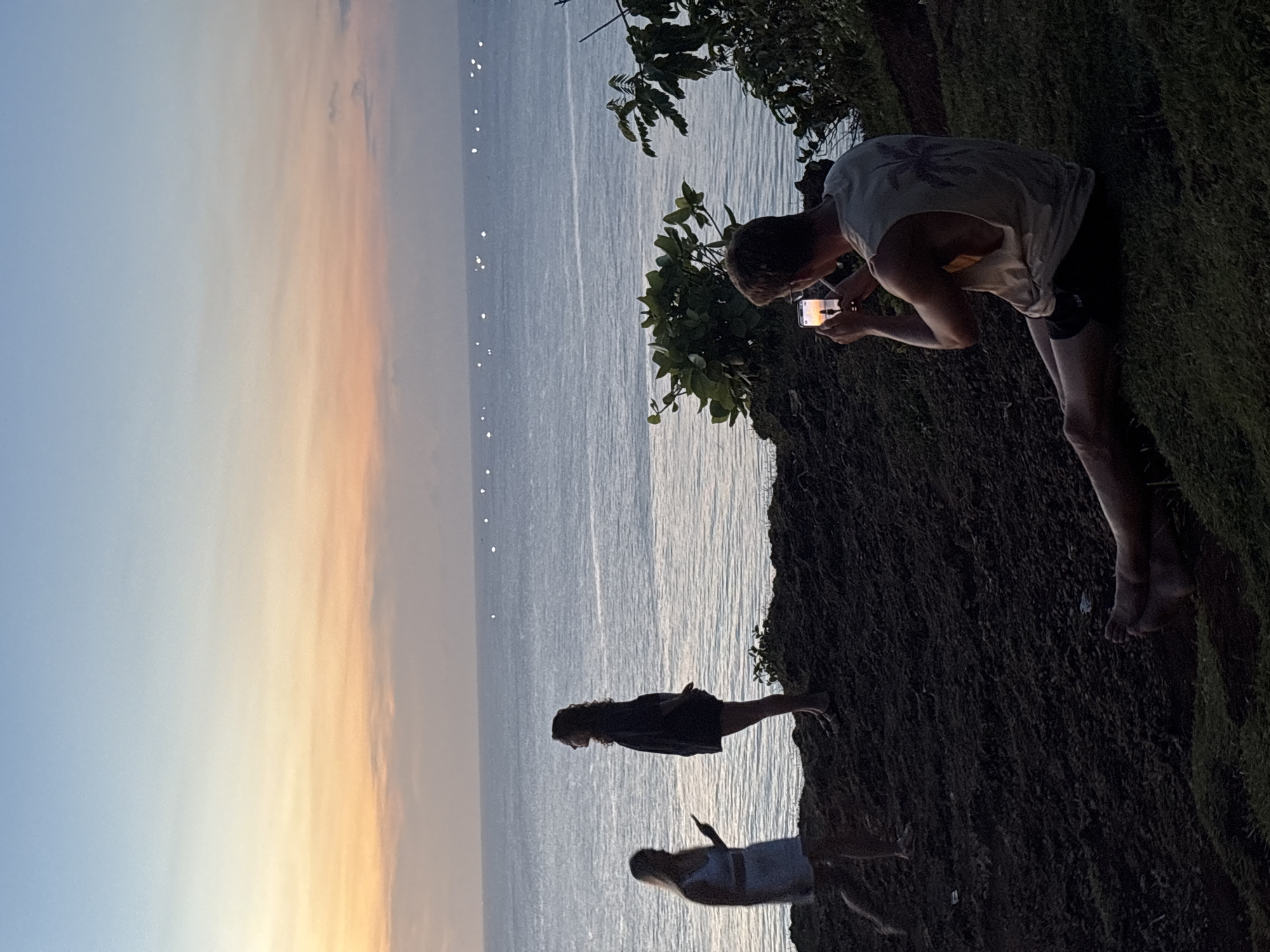 The image depicts a coastal scene in Kuta Selatan, Indonesia, at sunset. The sky transitions from blue to orange. The ocean is visible, dotted with small white lights. A man stands on a grassy cliff edge, using a smartphone to take a picture of the sunset. Below him, two women stand near the cliff's edge, facing the ocean.