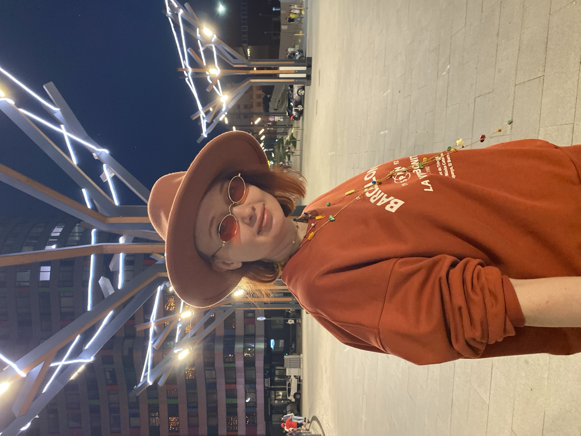 The image shows a person wearing a brown hat and red sunglasses, standing outside at night. The person is wearing a red-orange sweatshirt with the word "BARCA" printed on it. There are lights and building in the background. The location appears to be the city of Yekaterinburg, Russia.