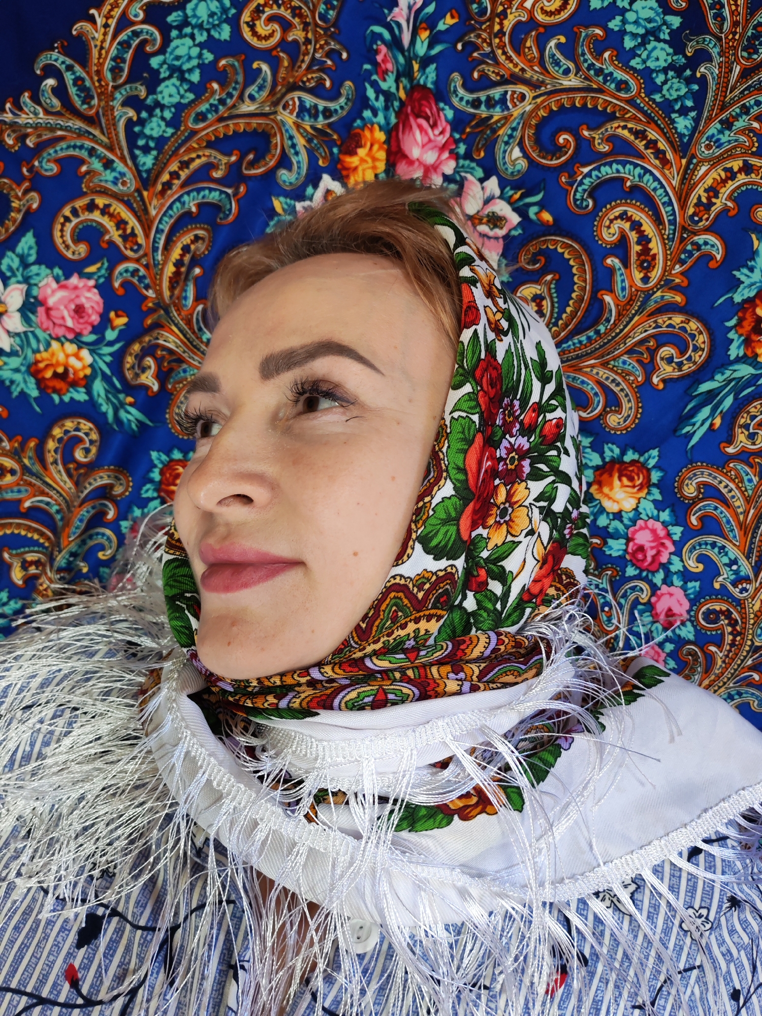 A woman with fair skin and reddish-brown hair, covered by a floral headscarf, looks off to the left. The headscarf features a predominantly white background with vibrant floral patterns in red, green, and yellow. She wears a white garment adorned with a fringe and a hint of blue and white striped material underneath. Behind her is a bright blue patterned background with floral and paisley designs in orange, yellow, and pink. The location is Chaikovsky, Russia.