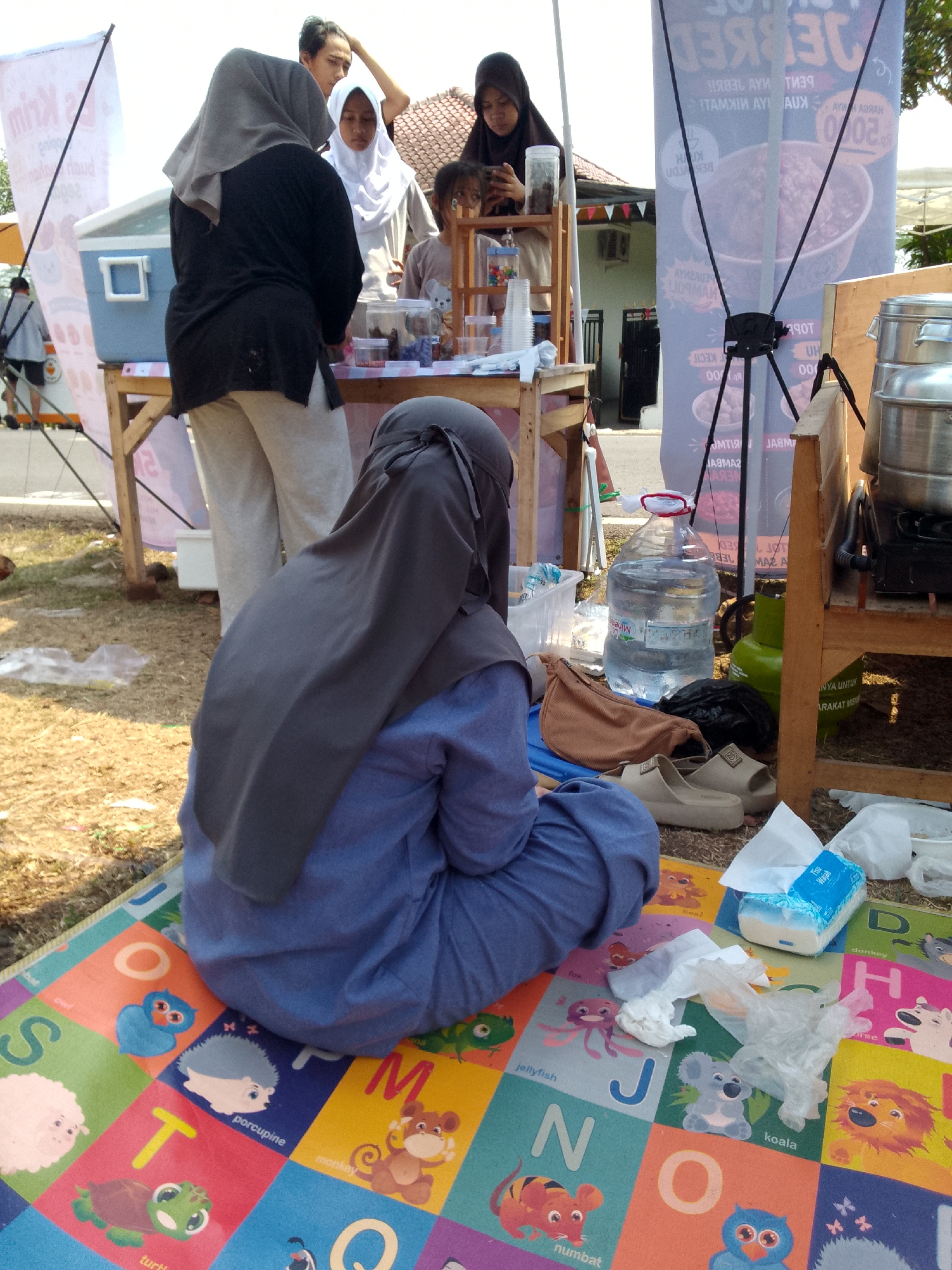 In Mandalawangi, Indonesia, a scene unfolds outdoors with a focus on a food stall or event setup. In the foreground, a person wearing a dark blue outfit and a grey hijab sits on a colorful alphabet mat, partially obscuring a large bottle of water, a brown fanny pack, and a pair of tan sandals. To the left, a blue cooler is visible on a wooden table laden with various items, including clear cups and containers filled with food. Several individuals are present in the mid-ground, including a woman in a white hijab and a woman in a black hijab, both interacting with the setup. A younger person is also visible, seemingly engaged with a phone. Behind them, a large banner with text and images is erected. The environment suggests a casual outdoor gathering or market, with dry grass and sunlight indicating warm weather. The overall impression is one of a community event or street food vendor operation.