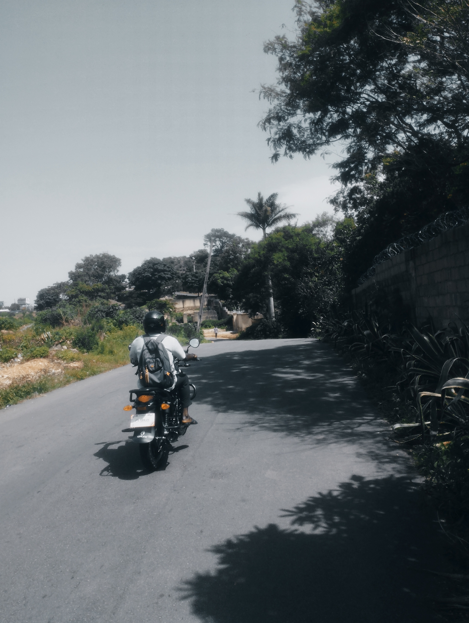 The image shows a person riding a black motorcycle on a paved road. The rider is wearing a black helmet and a grey backpack. The road is surrounded by lush green vegetation, trees, and a stone wall topped with barbed wire on the right side. The sky is overcast. The overall scene suggests an outdoor environment, likely in a rural or suburban area. Given the context provided, this scene could be located in the city of Jos, Nigeria, known for its hilly terrain and vegetation.