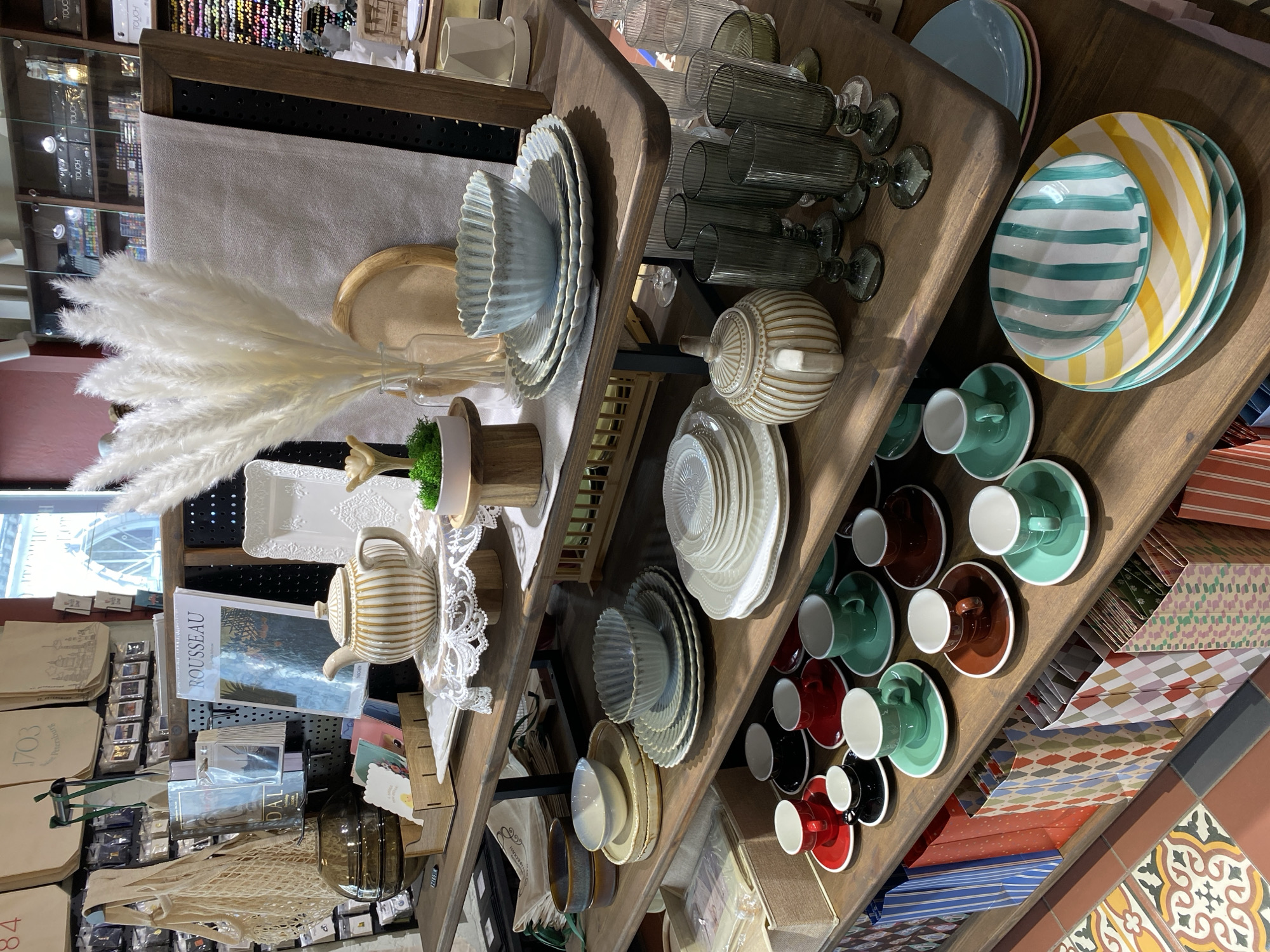 The image captures a vibrant, tilted view of a retail display, likely within a homeware or gift shop in Saint Petersburg, Russia. No people are visible, but the scene is filled with a meticulously arranged assortment of consumer goods.
On the left side of the frame, a large bouquet of white pampas grass serves as a central decorative element. Behind it, on a wooden shelf, are stacks of light blue ceramic plates and bowls with ridged, scalloped edges. Further right, a collection of smoky green fluted glass tumblers and goblets is displayed. Below these, a decorative teapot and a lidded sugar bowl with horizontal stripes in beige and white are prominent. Several white ceramic dishes with embossed patterns are also on display.
Toward the lower left, various books are visible, including one with "ROUSSEAU" on its spine and another with "DAL." Below these, fabric tote bags are stacked, some bearing the text "1703" and "St. Petersburg," explicitly linking the scene to the specified location.
The right side of the image showcases colorful dinnerware. Stacks of plates feature bold stripes in yellow and white, and teal and white. Below these, numerous teacups and saucers in vibrant teal, deep red, dark brown, and black are neatly arranged on wooden shelves. The very bottom right reveals a tiled floor with intricate, colorful geometric patterns. In the background, behind the main display, shelves filled with what appear to be art supplies or markers, with the word "TOUCH" visible on some boxes, are faintly seen through glass.
The overall impression is that of a thoughtfully curated store interior, presenting a variety of items for sale in an inviting manner. The lighting suggests an indoor setting, and the items are presented clearly for browsing or purchase.
