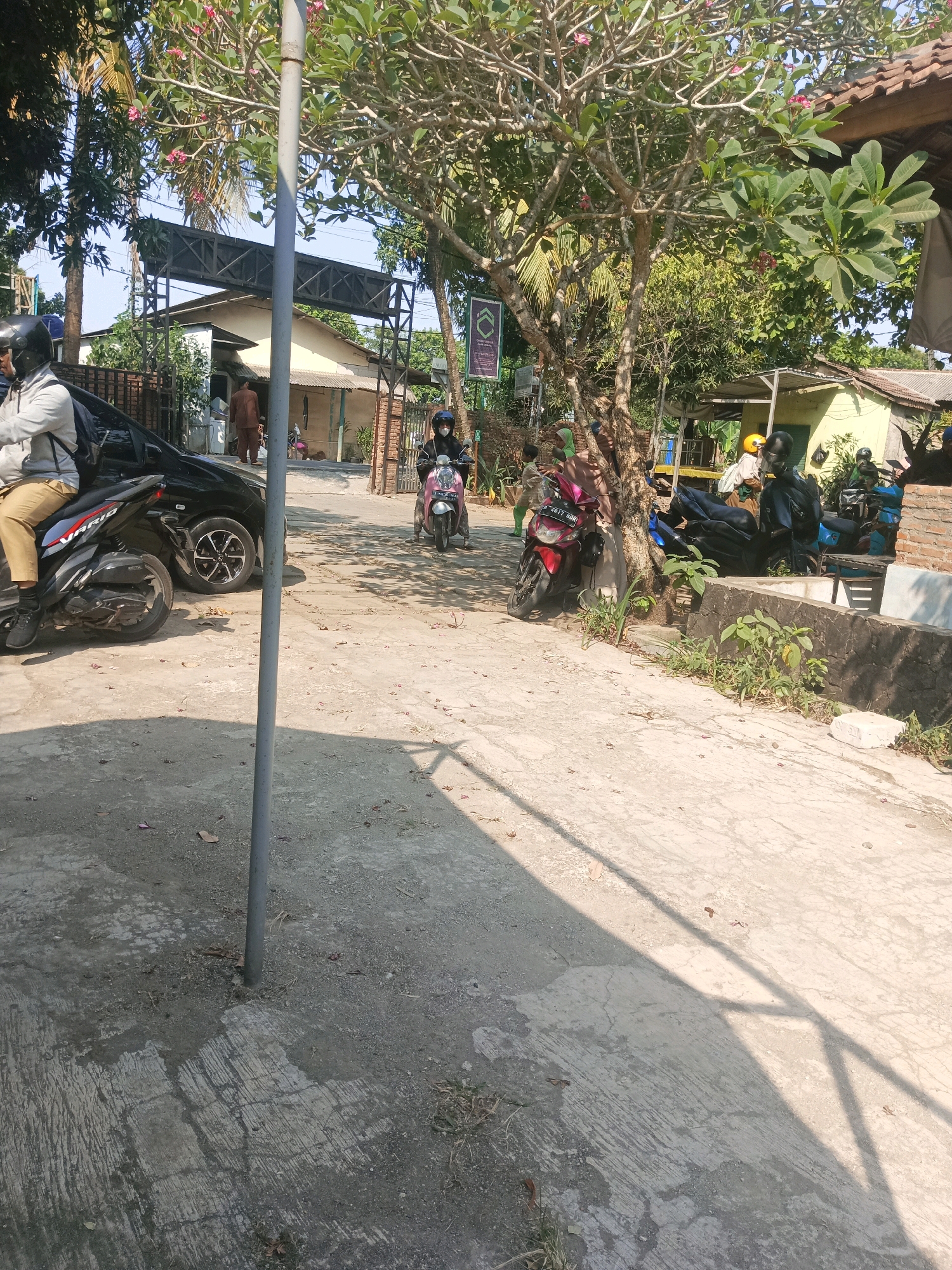 The image depicts a street scene in South Tangerang, Indonesia. A person on a motorcycle, wearing a helmet and a light-colored jacket and khaki pants, is partially visible on the left, next to a black car. Another person on a pink motorcycle, also wearing a helmet and a mask, is in the middle of the frame. A red motorcycle is parked on the right side of the road. Several other motorcycles are parked in the background, along with a building and trees. The ground is paved with concrete, and shadows from trees and structures are cast across it. The scene appears to be outdoors during the daytime, with clear skies.