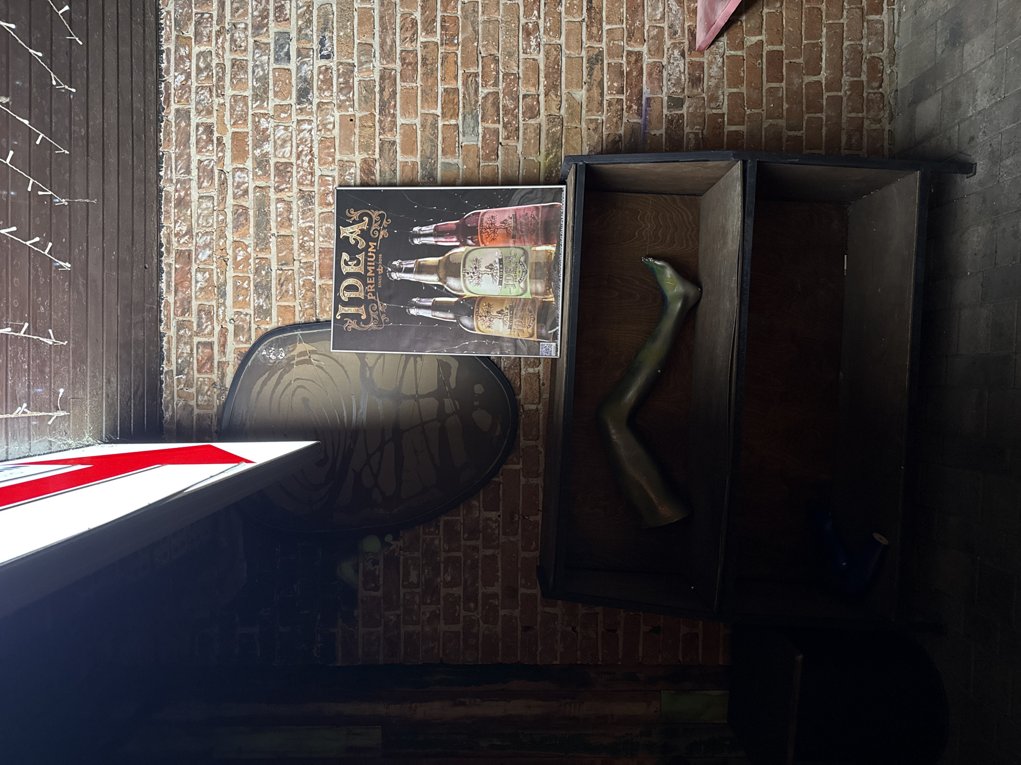 The image depicts an indoor scene with a brick wall as the background. A large poster advertising "IDEA PREMIUM" cider, featuring three bottles of the beverage, is prominently displayed on the wall. In front of the poster, a dark wooden shelf unit is visible. On the top shelf of this unit rests a mannequin's leg, posed at an angle. The bottom shelf is mostly in shadow, but a blue object, possibly a spray can or a bottle, is discernible. Above the brick wall, strings of small white lights are strung across the ceiling. To the left of the shelf unit, a part of a sign with red and white lettering is visible, casting a bright light downwards. The floor appears to be tiled with a grey pattern. The overall atmosphere suggests a bar or a retail space.