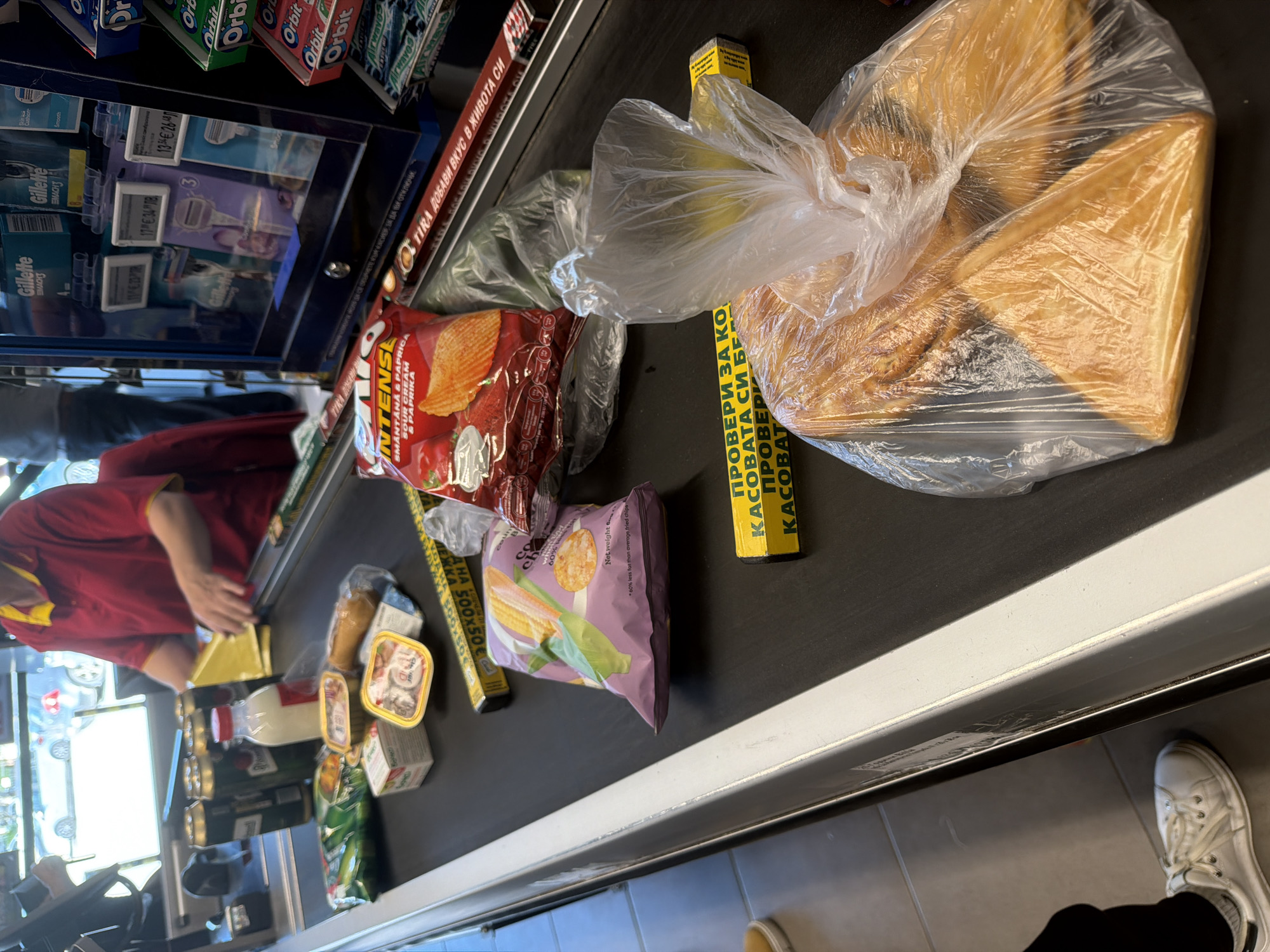 The image shows a checkout counter at a supermarket, likely in Plovdiv, Bulgaria, given the Bulgarian text on the checkout divider and packaging. A cashier in a red uniform is visible, processing groceries on the conveyor belt. Various items are on the belt, including: a bag of bread, bags of chips (including flavors like sour cream and paprika), canned goods, bottles, and other packaged food items. To the right of the checkout counter, a person in black pants and white shoes is partially visible. Behind the conveyor belt are shelves with personal care products like Gillette razors and Orbit chewing gum. The floor is tiled. The lighting indicates an indoor retail environment.