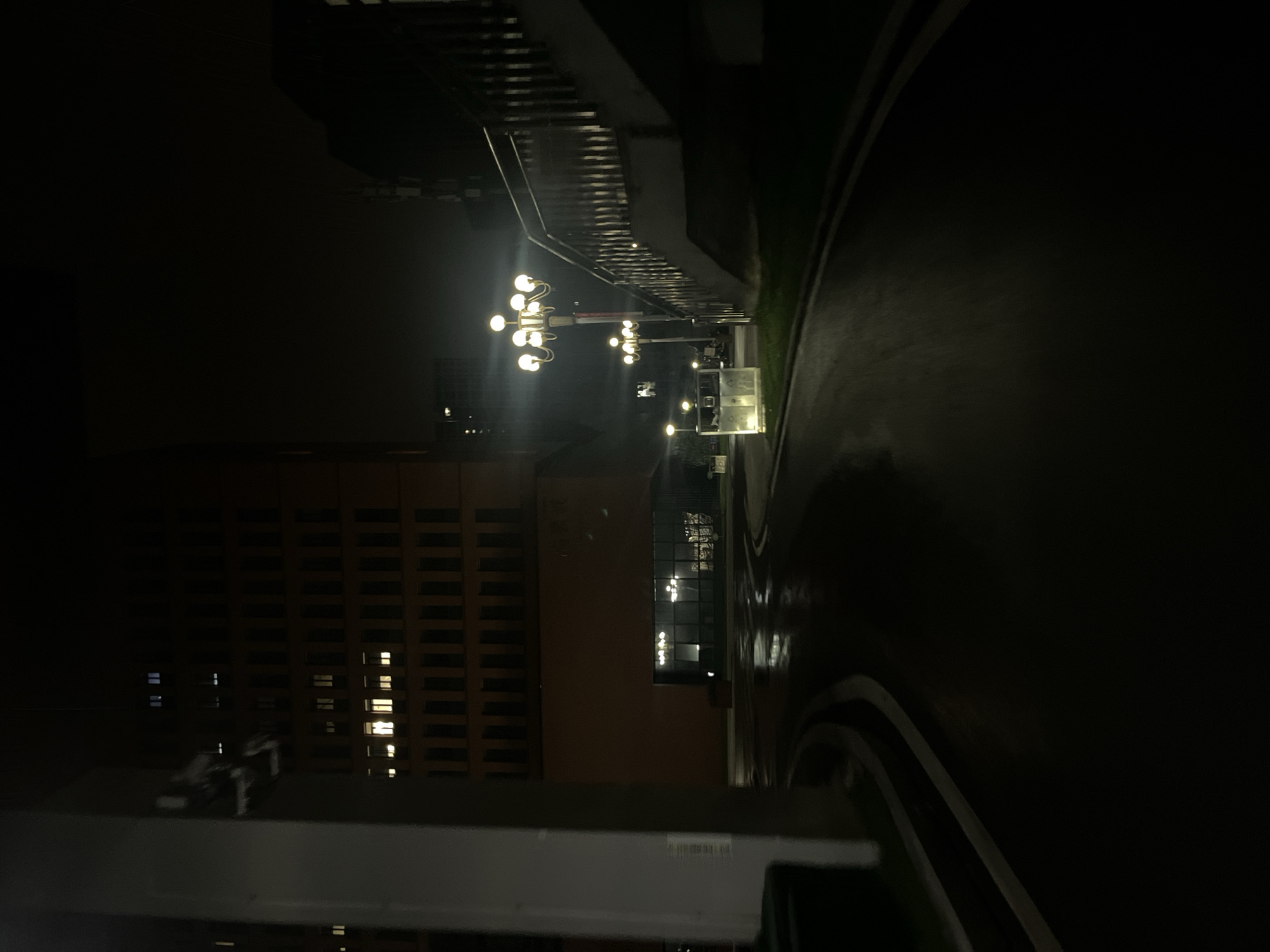 The image displays a nocturnal urban street scene in Yuzhong District, China, characterized by wet ground and artificial illumination. A curved, asphalt path with distinct white markings occupies the lower and central portions of the frame, reflecting light sources. On the left, a prominent light-colored, vertical support pillar stands in the foreground, partially obscuring a multi-story building with a reddish-brown facade. This building features numerous rectangular windows, several of which are illuminated with warm yellow light on various floors. At its base, a section with a glass exterior reveals an illuminated interior space. On the right side of the frame, a concrete pedestrian overpass or bridge with metal railings ascends diagonally upwards. The area is lit by multiple ornate streetlights, each comprising several spherical bulbs emitting bright yellow light, positioned predominantly near the bridge and along the path's edge. Further along the path to the right, several light-colored, rectangular public bins are visible. The background consists of dark, indistinct urban structures against a uniformly dark sky. No individuals, vehicles, or specific actions are depicted.