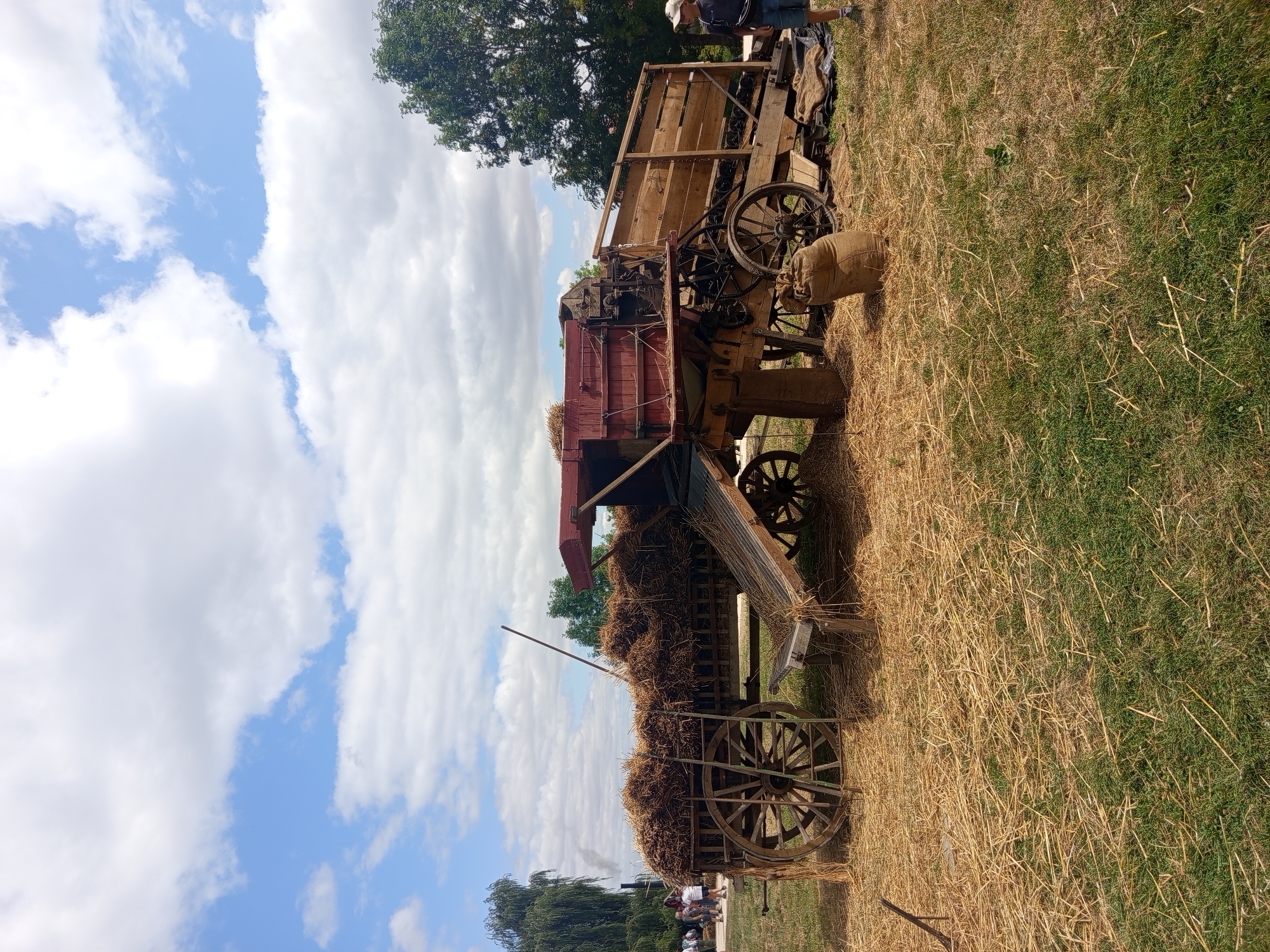 This image depicts an outdoor scene on a sunny day with a partly cloudy sky in Azannes-et-Soumazannes, France. The central focus is an antique threshing machine, a large piece of agricultural equipment, positioned in a field covered with dry grass and straw. The threshing machine is mounted on wooden wheels and appears to be made of wood and metal, with a red-topped structure. Hay is piled high on a wooden cart to the left of the threshing machine. In the background, several people are visible, some standing near a tall, weeping willow tree from which smoke is rising, suggesting a nearby building or activity. A person wearing a hat and shorts stands to the right of the threshing machine. Burlap sacks are placed on the ground near the equipment. The overall environment suggests a historical reenactment or demonstration of farming practices.