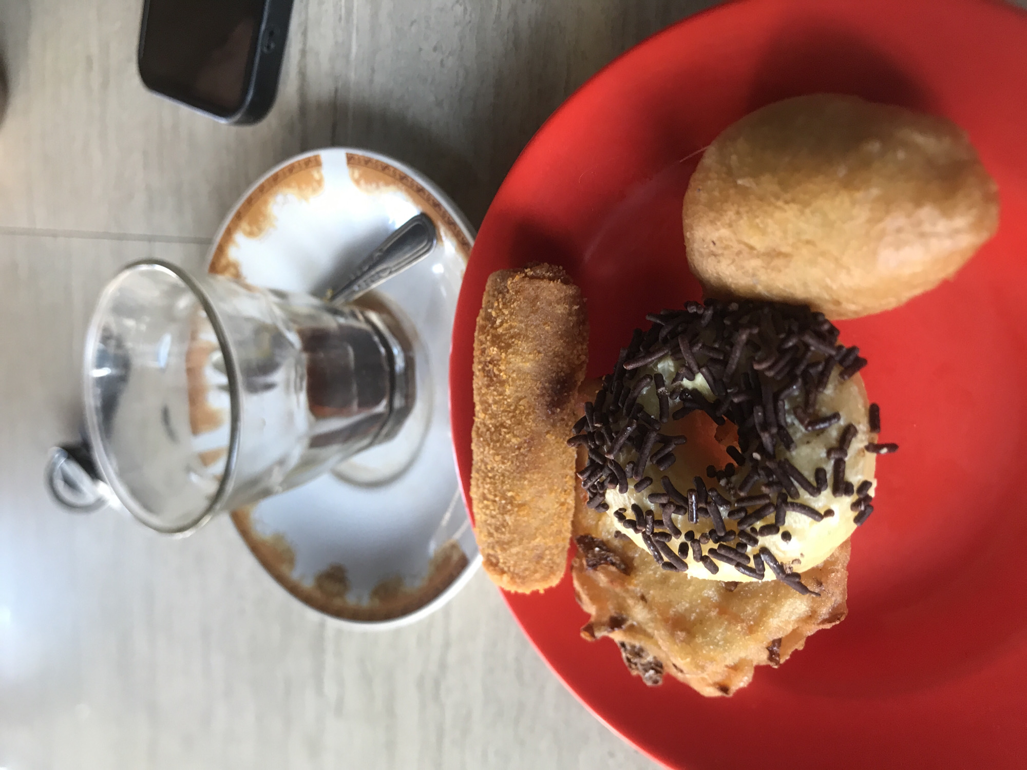 A red plate containing a donut with chocolate sprinkles, a small fried pastry, and a bun is positioned on a light-colored table. Beside the plate is a saucer with a spoon and a glass. A cell phone is visible in the upper left corner of the image. The pastries and tableware suggest this image was taken in a café or restaurant in Lhokseumawe, Indonesia, however, no additional contextual elements about the city or the scene are discernable. No people are visible.