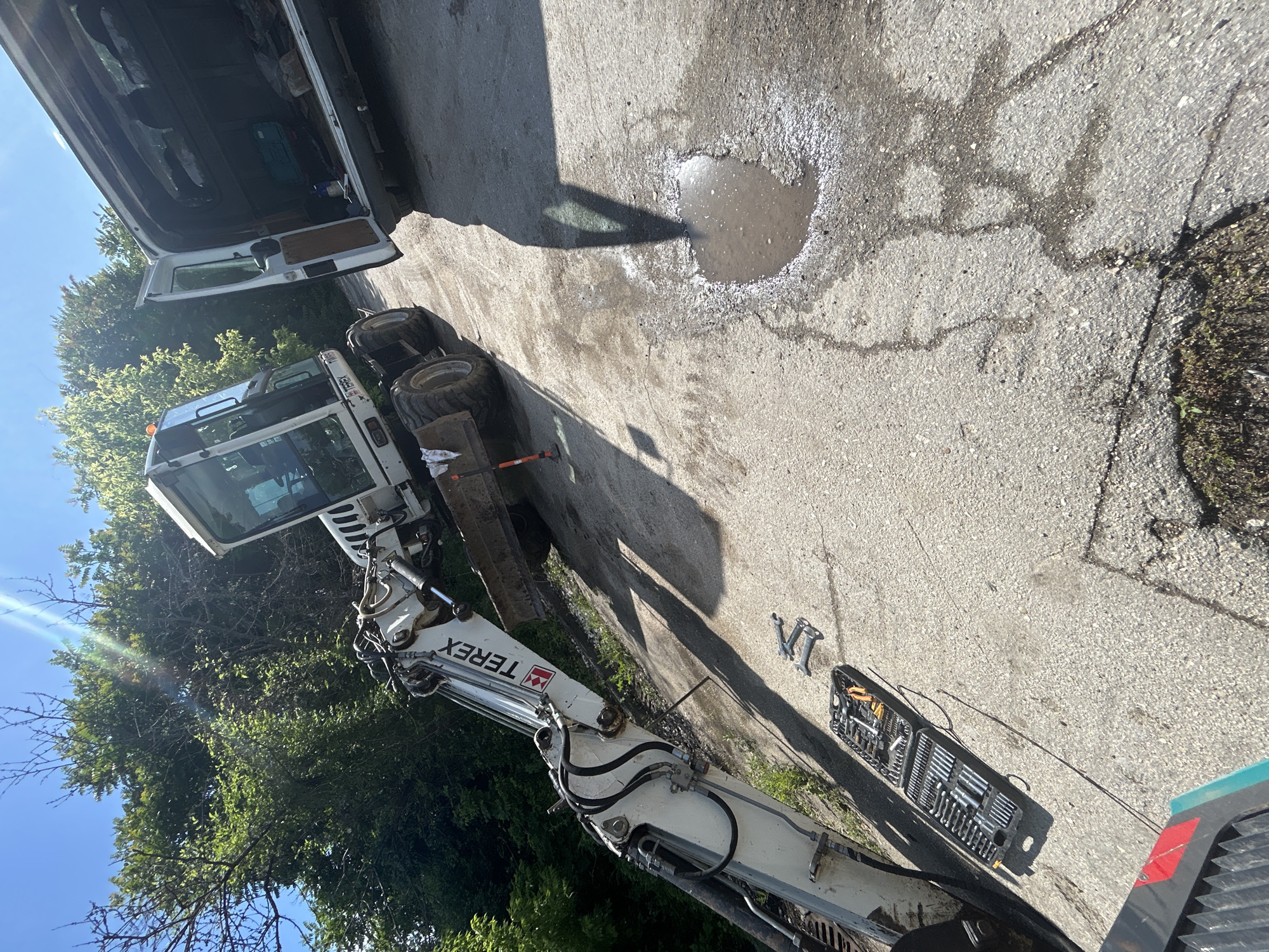 The image shows a construction site in what appears to be Silistra, Bulgaria, based on the Cyrillic lettering on the Terex excavator. A white van with its back door open is parked next to a Terex excavator. A puddle of water sits on the cracked concrete pavement. A set of tools is laid out beside the excavator. There are trees in the background and a blue sky.