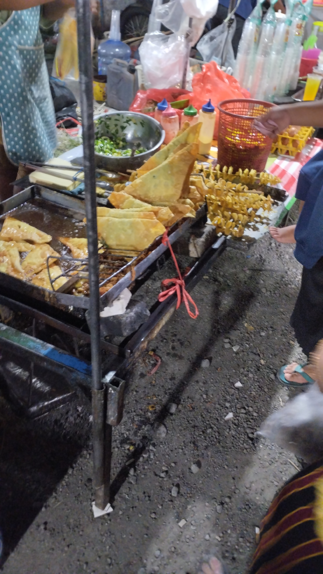 This media file depicts an outdoor food stall in Sambungrejo, Indonesia, likely in the evening or at night due to the artificial lighting and shadows. The scene is busy with activity. In the foreground, a food vendor is preparing fried snacks. A large metal tray filled with oil is actively frying golden-brown pastries, some of which are triangular in shape, resembling samosas or pastels. Tongs are placed inside the frying tray, suggesting ongoing cooking. Next to the frying tray, there is a bowl of chopped green vegetables, likely an ingredient or a side dish.
Behind the frying area, several condiments are visible, including bottles of sauce and a dispenser, suggesting options for customers. A red plastic basket is also present, and a hand is reaching towards it, possibly a customer or the vendor. Further back, there are stacks of spiral-cut fried potatoes on skewers, adding to the variety of food offered. In the background, various items are loosely arranged, including plastic bags, bottles of water, and what appears to be a motorcycle wheel, indicating a casual, street-side setting.
Two individuals are partially visible. One person, wearing a blue top and dark pants, is standing on the right side of the frame, wearing flip-flops. Another person, wearing a patterned garment with stripes of yellow, red, and black, is visible at the bottom right, also wearing flip-flops. The ground is covered in gravel and small stones, typical of an outdoor market or street environment. The overall atmosphere appears lively and commercial, with a focus on street food. No visible text is present in the image.