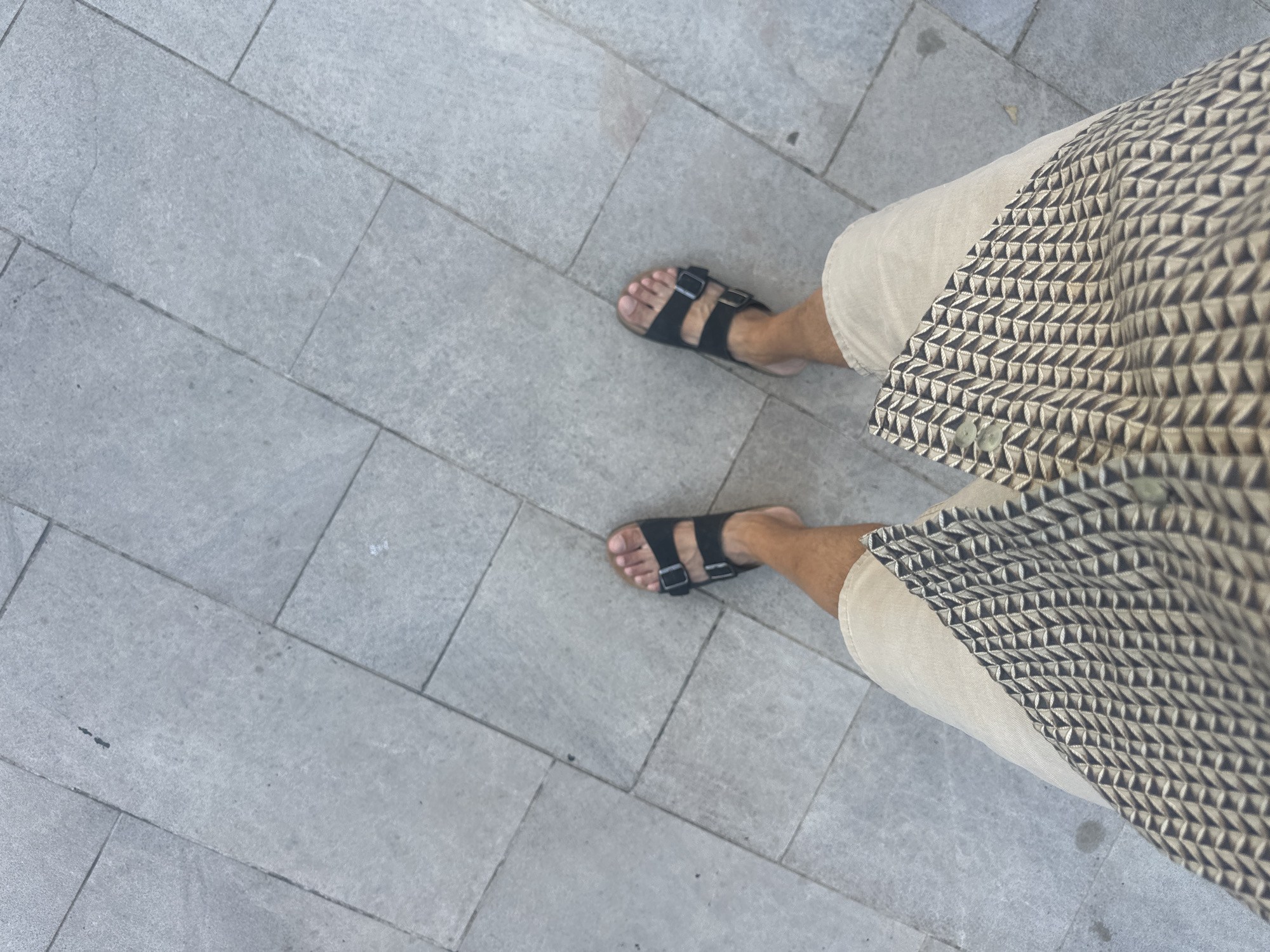 A high-angle, full-shot photograph shows a person's legs from the knees down, standing on a grey, tiled outdoor surface. The person is wearing tan-colored shorts and black Birkenstock sandals. The tiled surface appears to be part of a walkway or plaza. The image is composed in a way that emphasizes the texture and pattern of the tiles, as well as the geometric pattern on the person's shorts. The lighting suggests it is daytime, with a slight shadow cast by the person's feet. The context of Kumbor, Montenegro is not visually present in the image itself.