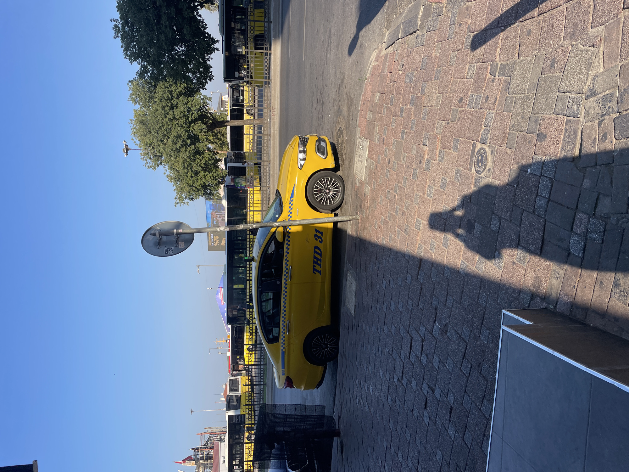 A yellow taxi with the license plate "THD 31" is parked on a cobblestone street in Kadıköy, Turkey. The taxi is positioned in front of a row of yellow buses. The scene is bathed in sunlight, casting a long shadow of a person, likely the photographer, on the ground. Trees and buildings are visible in the background, under a clear blue sky.