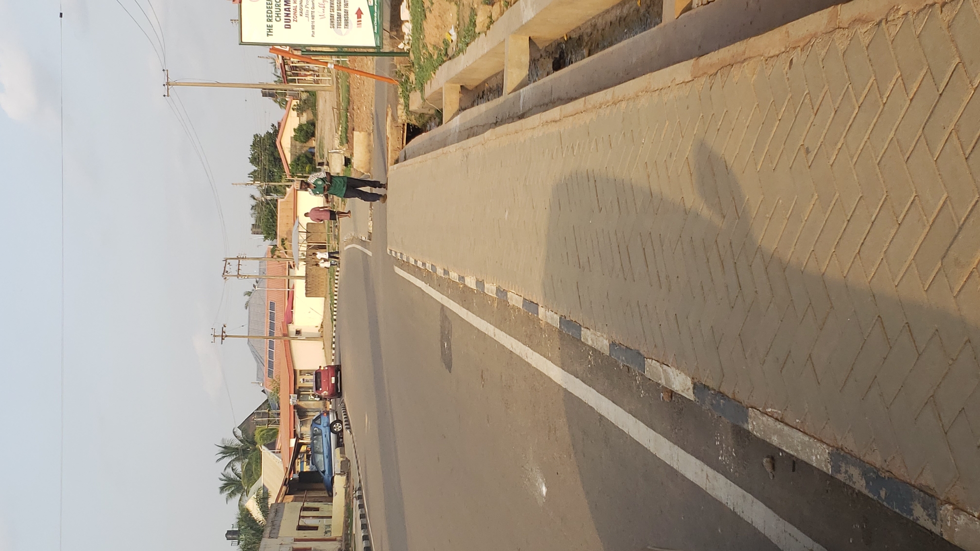 The image depicts an urban scene in Narayi, Nigeria, showing a street with two people walking on the side of the road next to a sign for a church. The road is paved and has a white line separating the lane closest to the sidewalk. The sidewalk is paved with herringbone patterned brick and bordered with black and white paint. A concrete barrier separates the sidewalk from a ditch. Beyond the road are buildings, some with solar panels, and utility poles with wires running overhead. A blue car and a red vehicle are parked on the side of the street. The sky is pale.