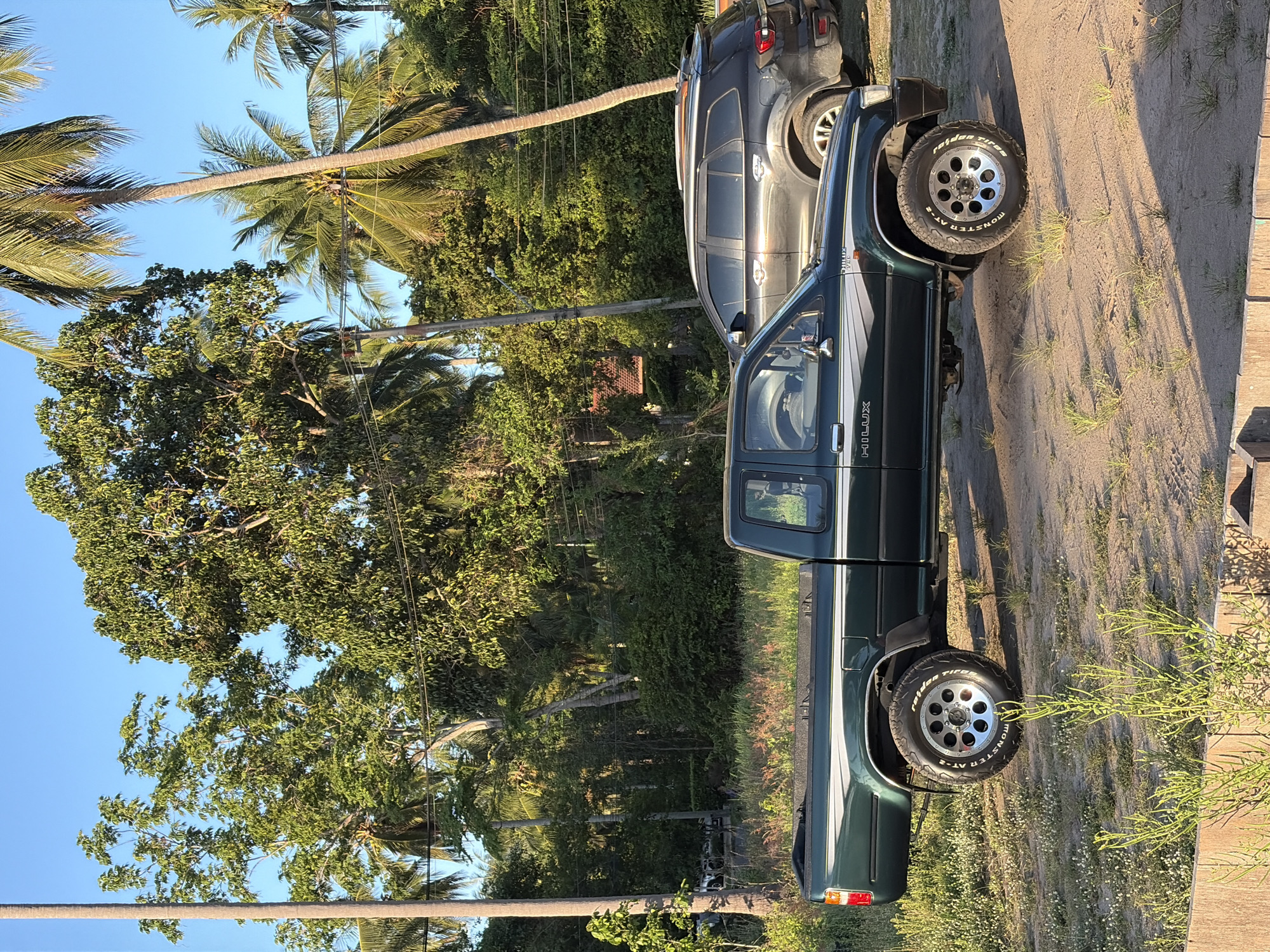 A dark green Toyota Hilux pickup truck is parked on a dirt lot. The truck is facing right and is in the foreground. The truck has a chrome trim along the side and a "HILUX" logo on the door. The truck also has off-road tires with "MONSTER TRUCK" written on the side. In the background, a dark gray SUV is parked to the right of the truck. Behind the vehicles, there is a dense jungle with palm trees and other greenery. The sky is clear and blue. The scene is bathed in the warm light of the late afternoon sun. The location is identified as Baan Taling Ngam, Thailand.