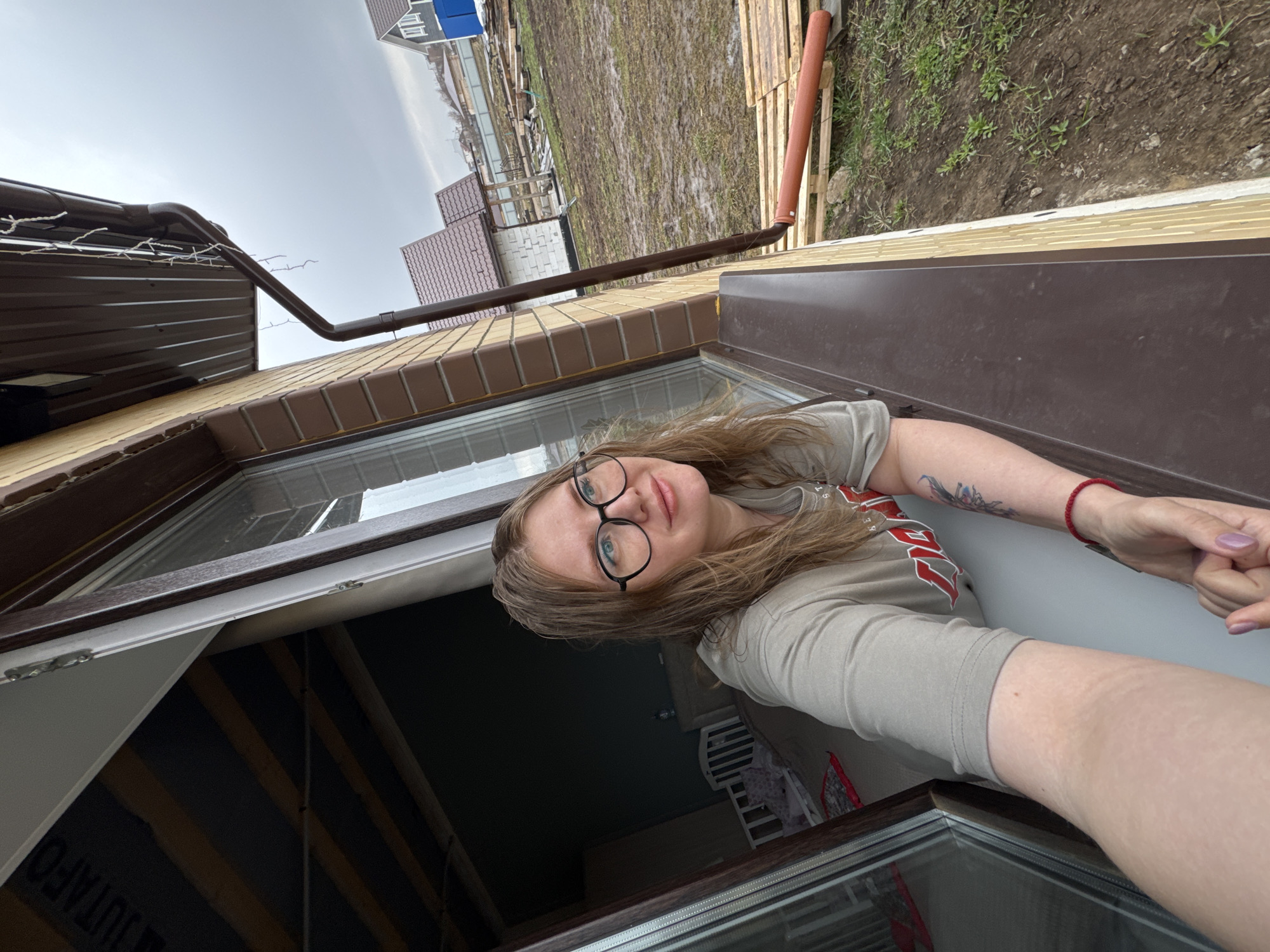 A woman takes a selfie, looking upwards from an open window or skylight of a building in Gornyi (Dvurechenskoe S/P), Russia. She has long brown hair, wears black-rimmed glasses, and has a neutral to slightly pleasant expression. She is wearing a light-colored t-shirt with a faint graphic design on the shoulder/sleeve, a red bracelet on her left wrist, and a tattoo on her right arm. Her fingernails are painted a light purplish-pink.
The scene outside the window reveals an overcast sky and damp, muddy ground with sparse patches of grass, suggesting cloudy weather or recent rain. The building features a brown gutter system, a section of light brown brickwork, and brown corrugated metal siding. In the background, other houses with brown roofs are visible, along with a building with a blue roof in the distance. A wooden pallet and an orange pipe are present on the ground beside the building. Inside the structure, visible through the window opening, are exposed wooden beams and dark insulation material with the text "JUTAFOL" faintly visible. The time of day appears to be daytime, under a uniformly gray, cloudy sky.