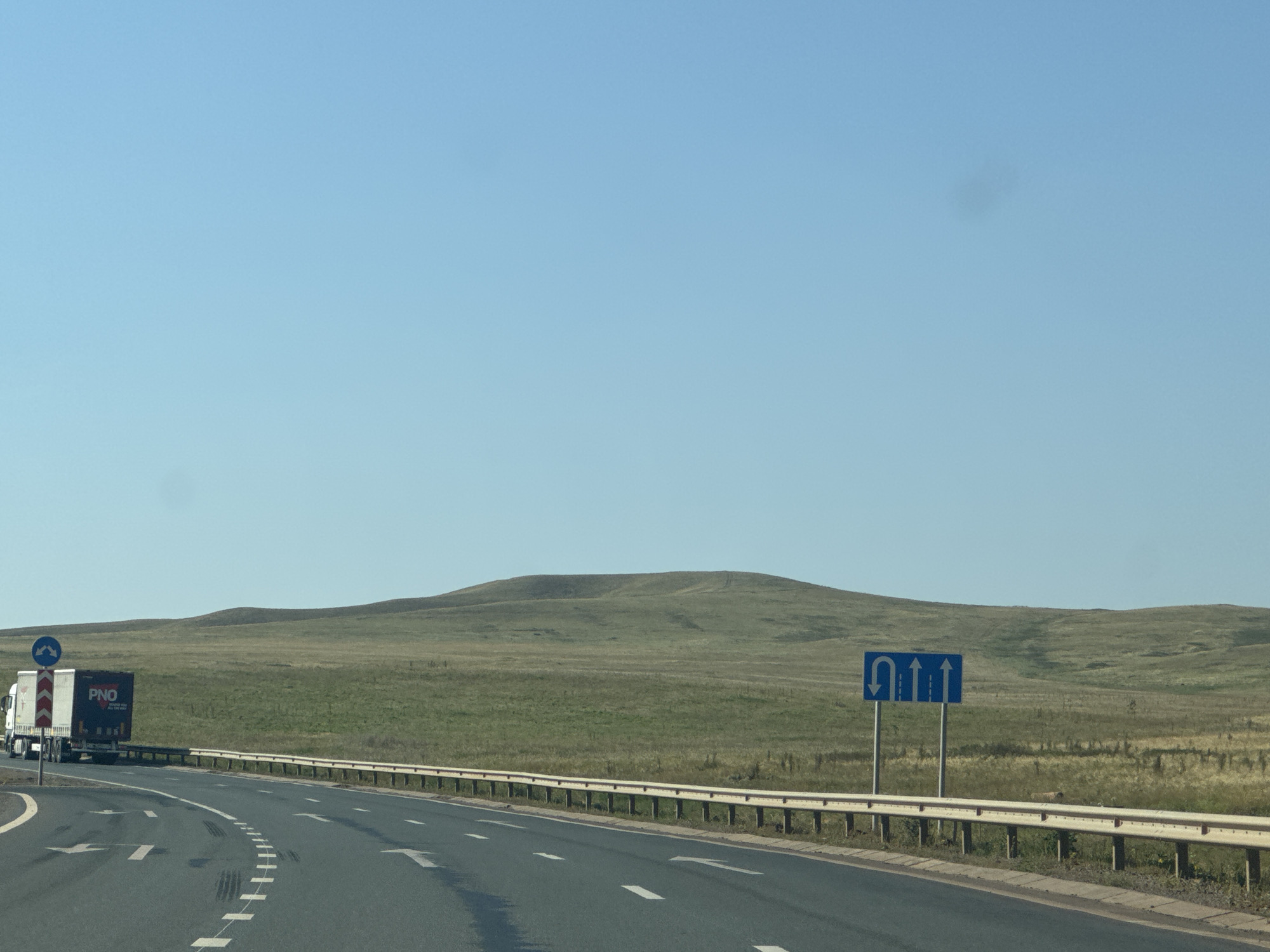 The image depicts a clear, sunny day with a bright blue sky. A paved road, marked with white lane lines and arrows, curves to the left. A guardrail runs along the right side of the road. On the left side of the road, a white truck with a dark blue trailer bearing the logo "PNO" is visible. A blue traffic sign indicating a U-turn and straight lanes is positioned on the right side of the road. In the background, rolling hills covered in dry grass stretch to the horizon. The landscape appears to be a rural area, possibly on the outskirts of Syrt, Russia. There are no people visible in the image.