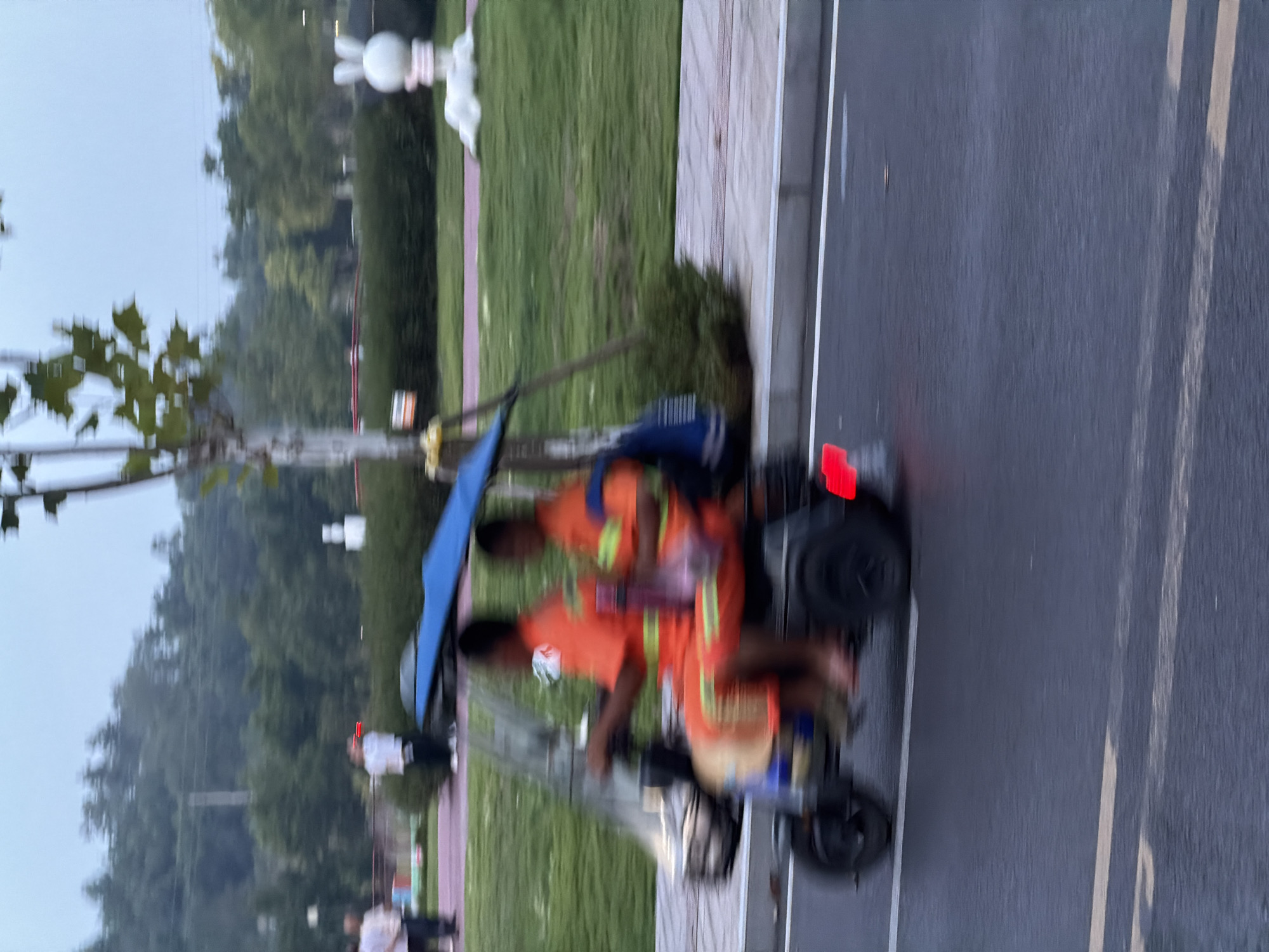 In Jiangjin, China, on a cloudy day, two individuals wearing bright orange uniforms are riding a three-wheeled electric vehicle on a wet asphalt road. The vehicle has a blue canopy for shade. The driver is on the left, and the passenger is on the right. Both are looking forward. The background shows a green grassy area with trees and a white rabbit sculpture. The image is blurred due to motion.