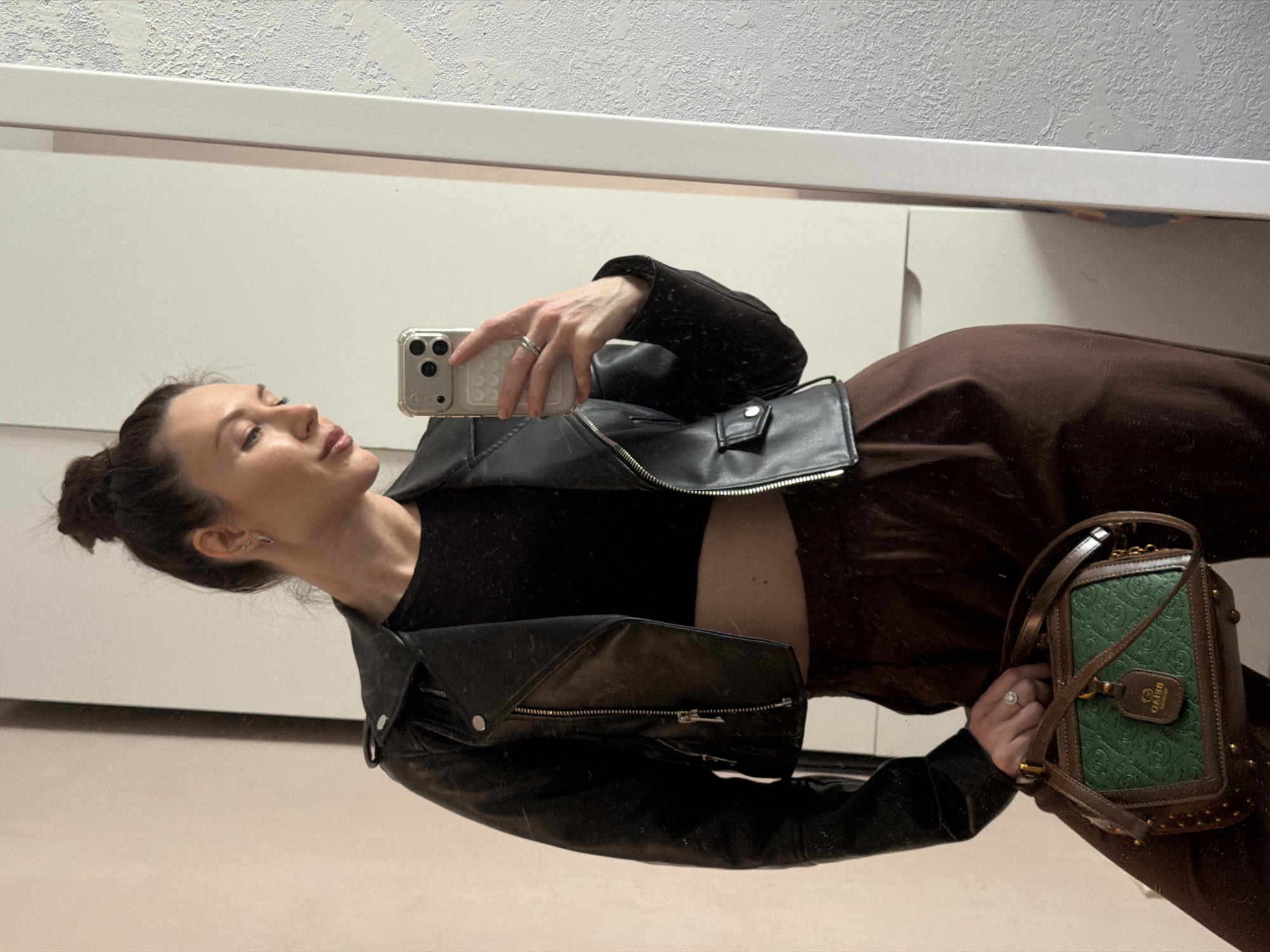 A young woman takes a selfie in a mirror. She wears a black leather jacket over a black top, brown pants, and holds a green and brown handbag. She has her hair up in a bun. The background shows a plain white wall and a mirror frame. The location is unknown.