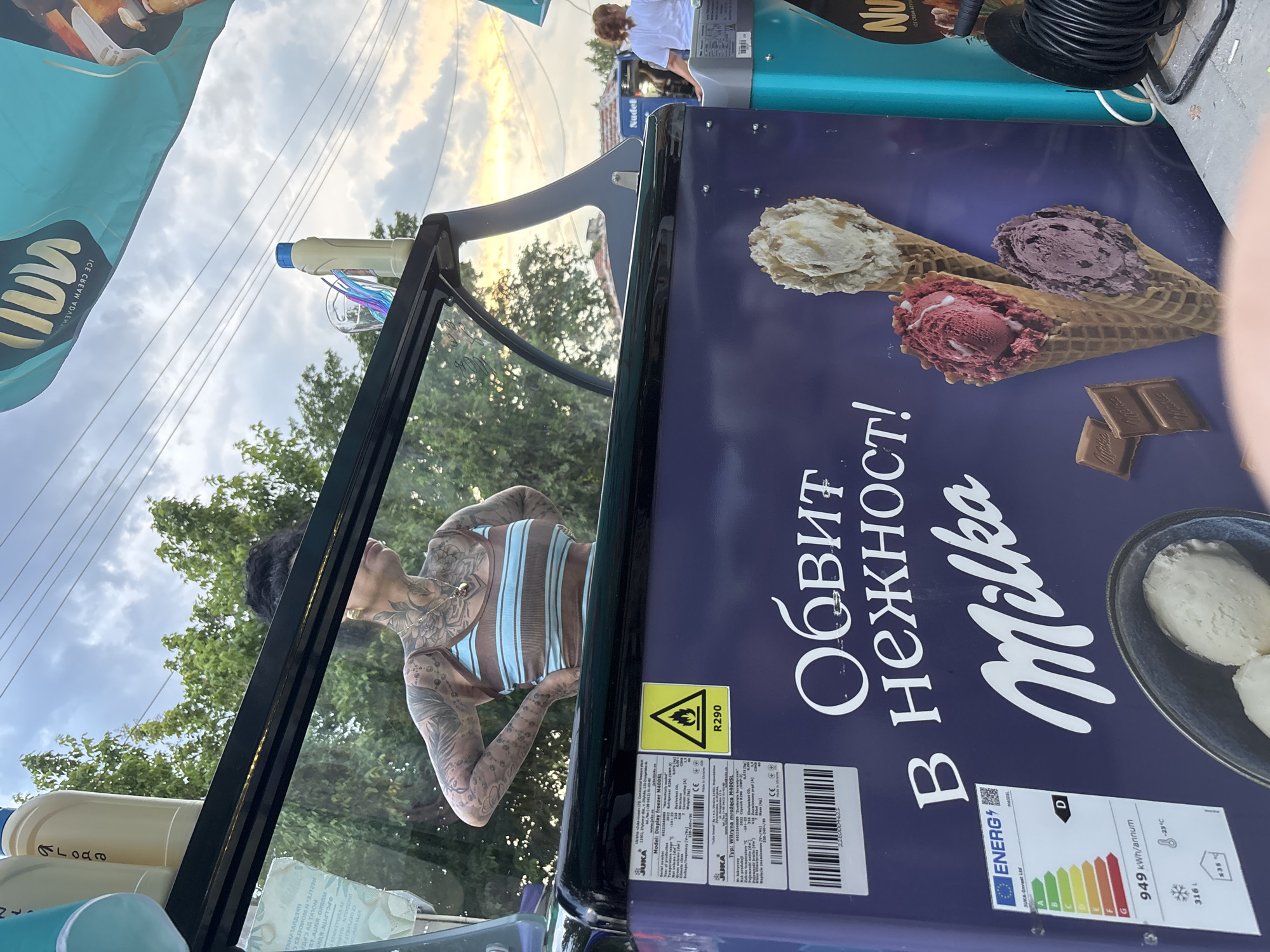 This image captures an outdoor scene at an ice cream stand in Kostievo, Bulgaria, during what appears to be late afternoon or early evening. The central focus is a Milka-branded ice cream freezer, primarily purple with white Cyrillic text reading "ОБВИТ В НЕЖНОСТ!" (Wrapped in tenderness!) and the "Milka" logo. The freezer displays images of various ice cream cones and scoops in a bowl, alongside Milka chocolate squares.
Affixed to the freezer are several labels: a technical data sheet from JUKA trade-Invest Ltd. for a "Display Freezer M400SL," indicating it is "Made in Ukraine" and uses R290 refrigerant, a flammable gas, as noted by a yellow warning sticker with a flame symbol. An energy efficiency label shows a "D" rating, 949 kWh/annum, and -23°C operating temperature.
Reflected in the glass top of the ice cream cart is a person, likely a woman, with dark hair and extensive tattoos covering her arms, shoulders, and chest. She wears a brown and light blue striped tank top and has her hands on her hips, facing to the right in profile. On the top surface of the cart, several items are visible, including light-colored bottles (one with a blue cap) and clear glasses containing colorful elements, possibly straws.
In the background, on the far right, a second person with reddish-brown hair tied back, wearing a white t-shirt, is partially visible, facing away from the camera. The setting includes green trees, overhead power lines, and a cloudy sky suggesting approaching dusk. Part of a light blue awning or flag, bearing reversed text that includes "ICE CREAM ADVENTURE," is visible in the upper right. A coiled black cable or hose is present in the lower right foreground. Distant structures, including one with "Nudel" visible on its side, are vaguely discernible.