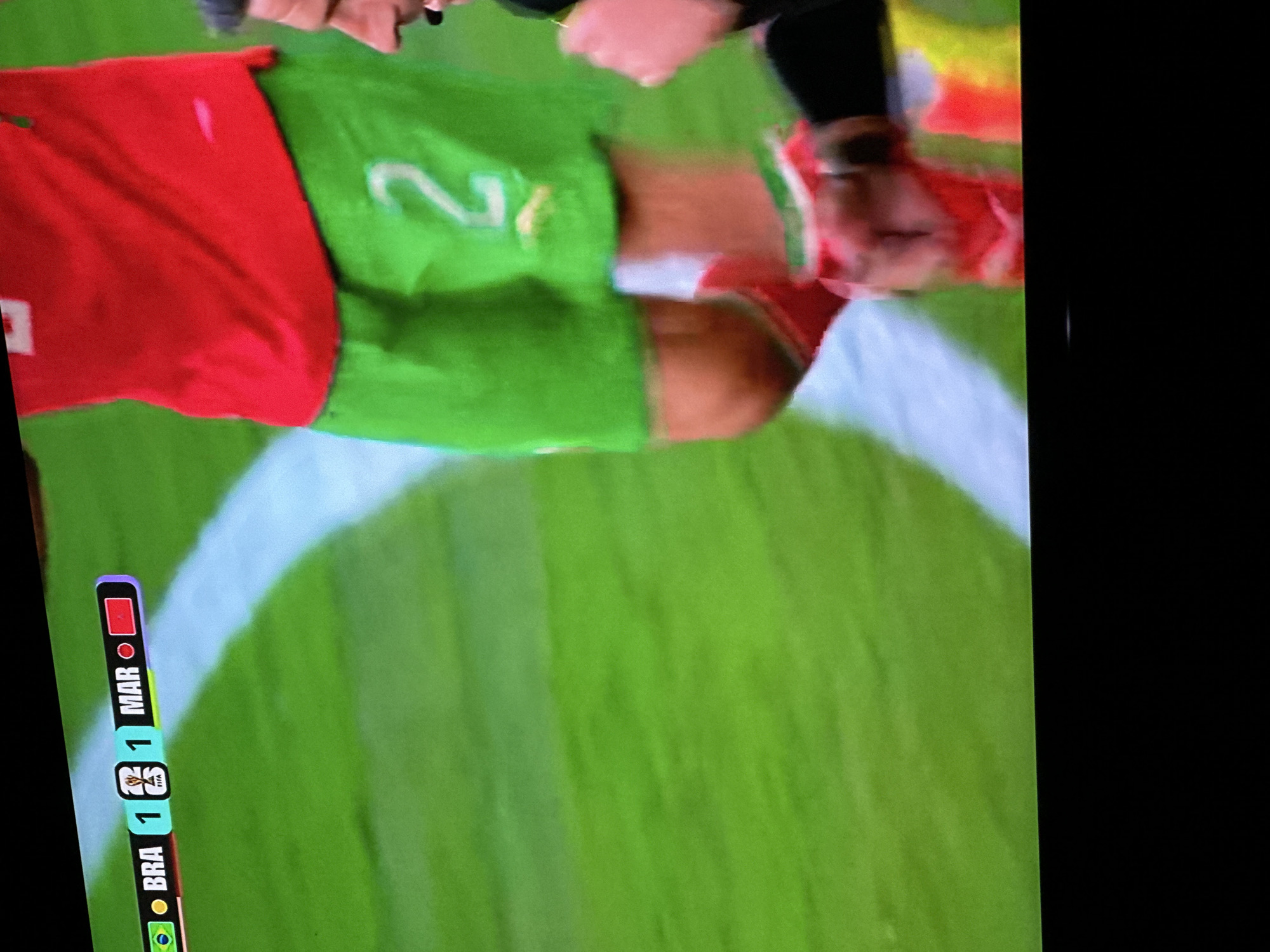 The image displays a television screen showing a soccer match scoreboard. The scoreboard indicates "BRA 1 1 MAR," with the Brazil flag represented by a green and yellow circle and the Morocco flag by a red rectangle. A player in a red jersey with green shorts, displaying the number "2" on their shorts, is visible on the right side of the screen. The environment appears to be a green soccer field with a white line. The television is placed in a dark room, with the bottom of the screen showing a black bar. The provided information about the city of Rayfield, Nigeria is not visually represented in the image and cannot be confirmed.