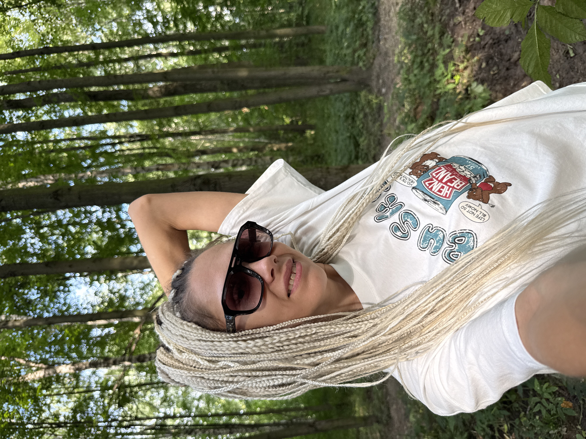 A woman with blonde braided hair and sunglasses is taking a selfie in a forest. She is wearing a white t-shirt with a cartoon image of Heinz baked beans on it. The background is a blur of green trees and foliage, suggesting an outdoor, natural setting. The lighting indicates it is daytime. The image quality is good, with a shallow depth of field that blurs the background. The woman's pose and expression are casual and relaxed. The location is specified as Rusanovo, Russia.