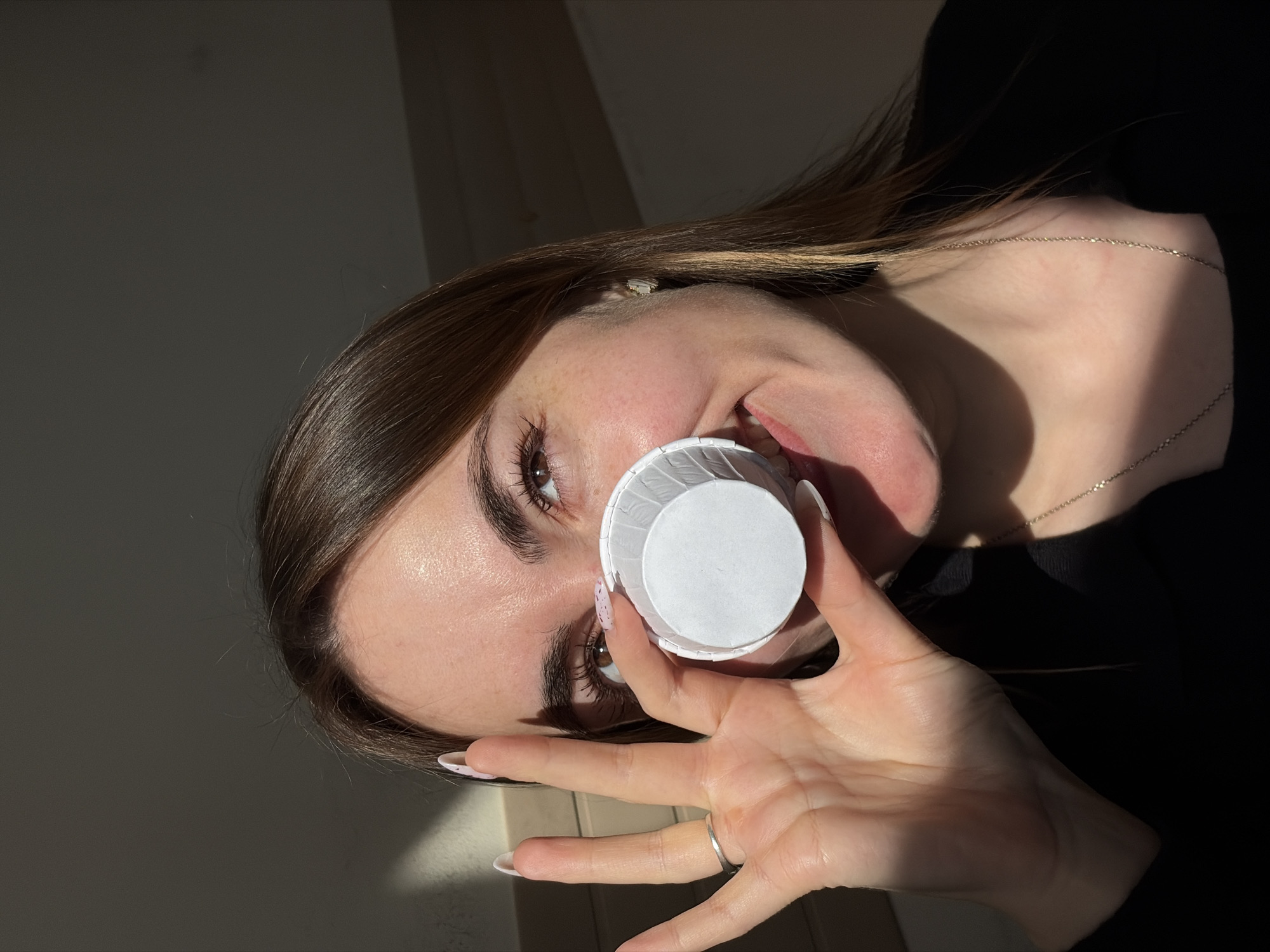 A young woman with light skin and brown hair is holding a small, white paper cup in front of her face. She is wearing a silver ring on her left hand and a delicate silver necklace. Her fingernails are long and painted light pink with speckles. The background consists of a white wall and a black garment. The lighting is dramatic, with strong highlights and shadows.
The location is not identifiable as Izhevsk, Russia based on the provided image.