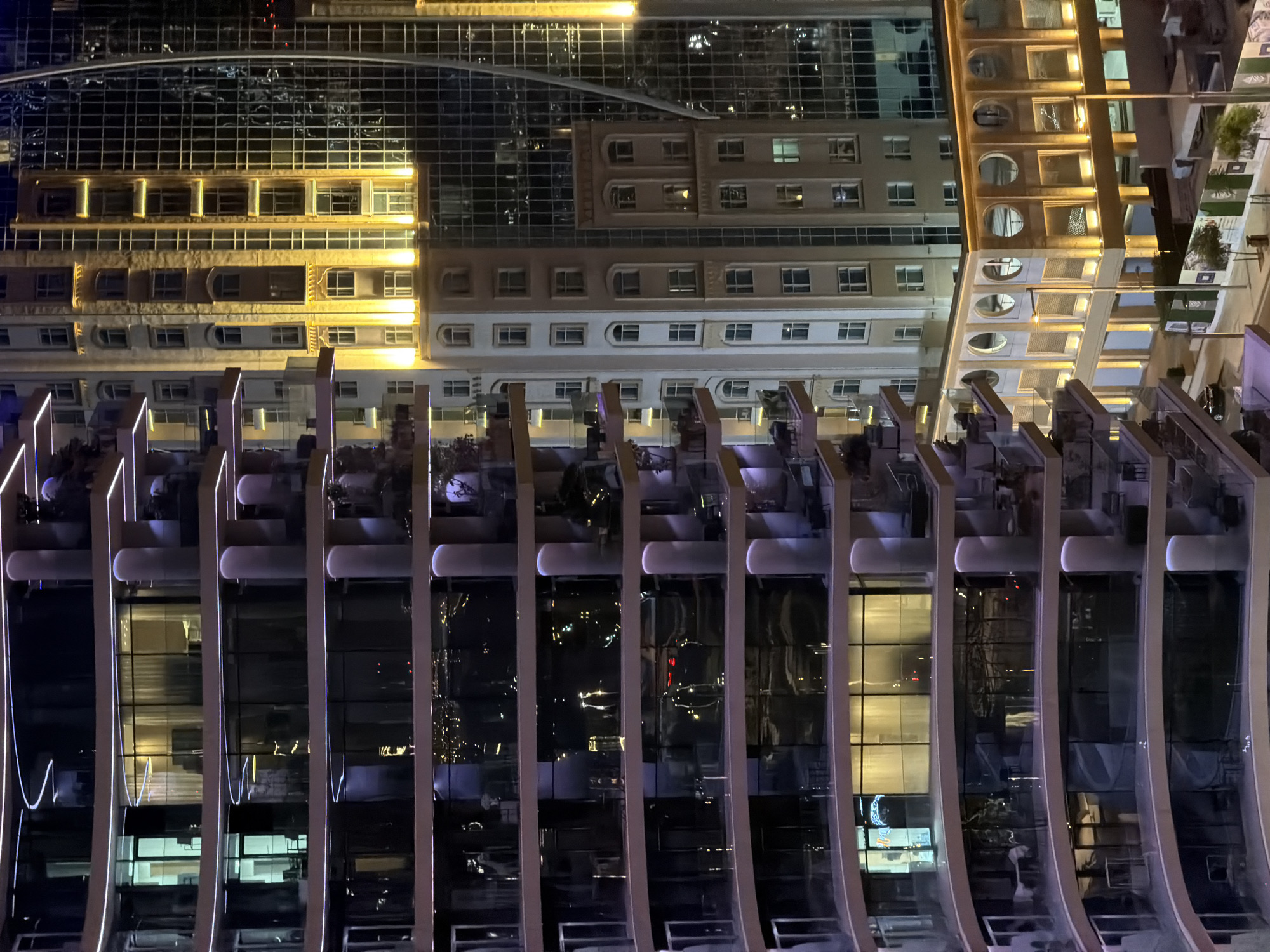 This nighttime image depicts an urban scene featuring a cluster of interconnected buildings, presenting a mix of architectural styles. On the left, a prominent modern high-rise building curves towards the viewer, distinguished by numerous balconies illuminated with continuous, glowing purple-white LED strip lights along their edges. These balconies feature glass railings and contain outdoor furniture such as chairs and small tables. Various interior lights are visible through the large glass windows, revealing glimpses of rooms. To the right of this modern structure stands a mid-rise building with a more traditional facade, characterized by multiple rectangular and arched windows and ornate decorative elements. This building is warmly illuminated by yellow uplights and downlights that accent its architectural details. Further to the right and partially obscured by the traditional building, a tall, contemporary glass skyscraper with a gridded facade reflects city lights. Some vertical warm yellow lighting is also visible on its surface. At the bottom of the image, the street level is visible, featuring a dark-colored car, streetlights, and a low-rise building with distinct circular windows, also warmly lit. Construction barriers or temporary fencing with green and white patterns line the street, alongside a few small trees. No individuals are discernible in the scene. The overall environment is a brightly lit city at night in Unknown, Unknown.