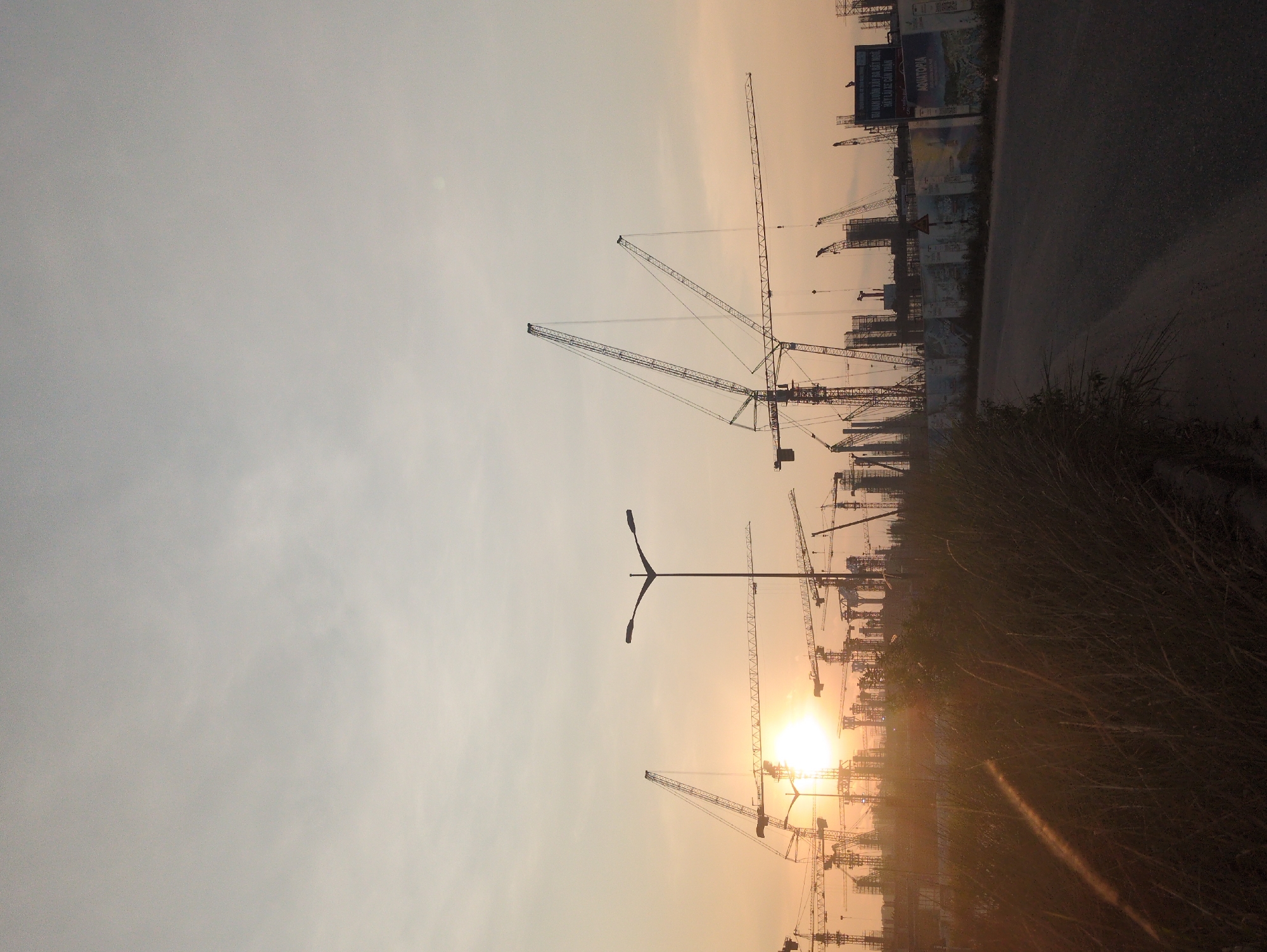 The image depicts a construction site in Phú Quốc, Vietnam, at sunrise or sunset. Several tower cranes are visible against a hazy, orange-tinted sky. A lamppost stands in the foreground. A road with signage and partially obscured structures runs along the right side of the image. Tall grass fills the lower right corner. The sun appears to be setting or rising behind the cranes, creating a silhouette effect.