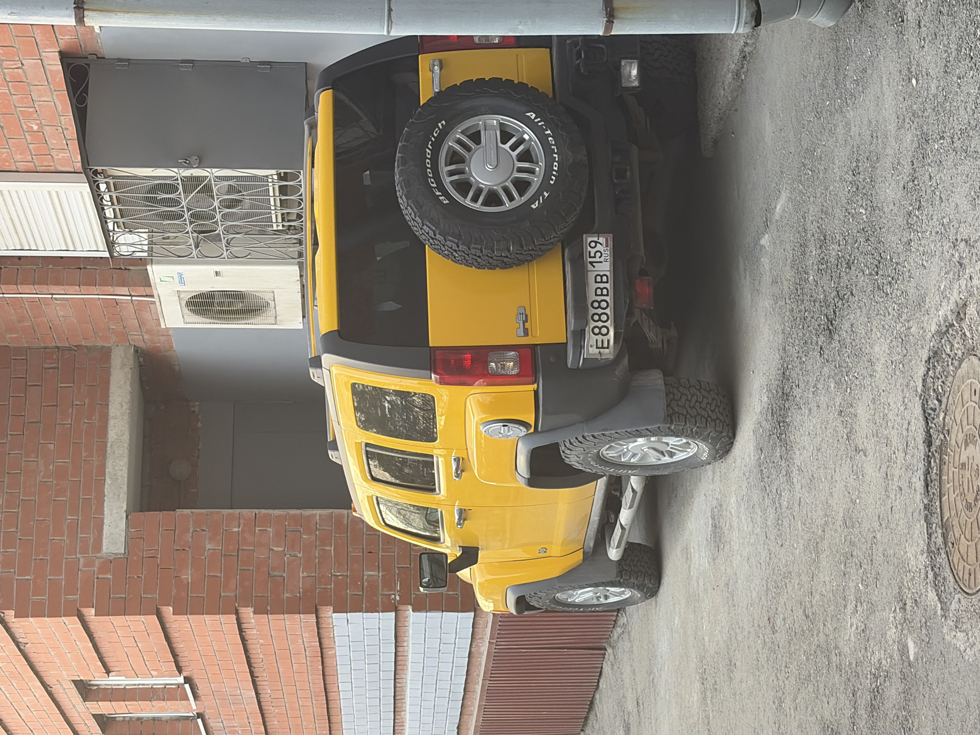 A yellow Hummer H3 with license plate E888BB 159 RUS (indicating Perm, Russia) is parked on a concrete surface. The vehicle is positioned between a brick building on the left and a corrugated metal fence on the right. A manhole cover is visible on the right side of the image. An air conditioning unit and a caged window are attached to the building. A horizontal pipe runs across the top of the image.