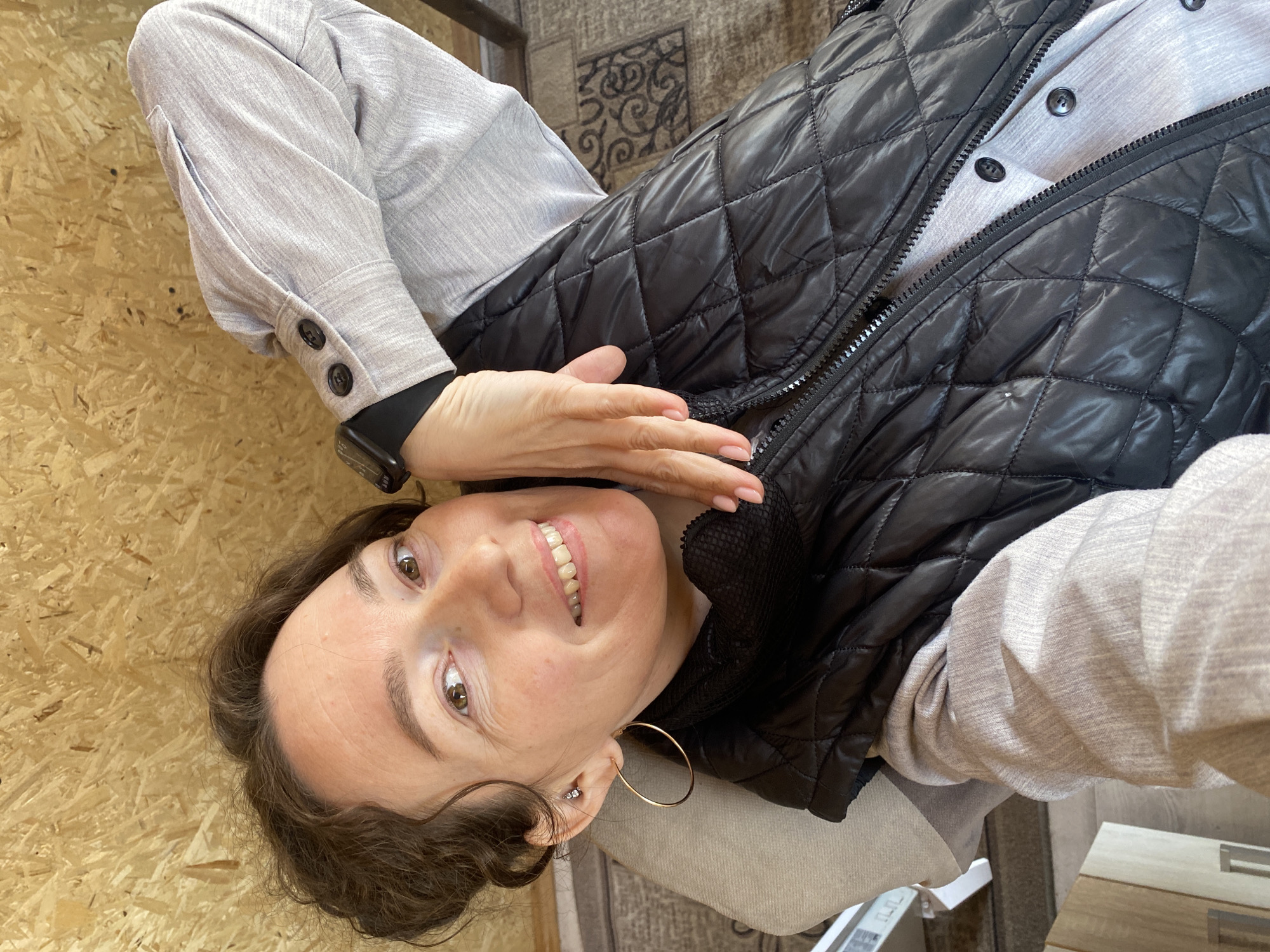 A woman is lying down, smiling, and looking at the camera. She is wearing a light gray long-sleeved button-down shirt with a black quilted vest over it. She has brown hair and is wearing gold hoop earrings and a black smartwatch. Her right hand is near her face.
The background appears to be a light-colored wooden wall on one side and a patterned rug on the other. There is no discernible information to confirm the location as Ol'khovskoye, Russia.