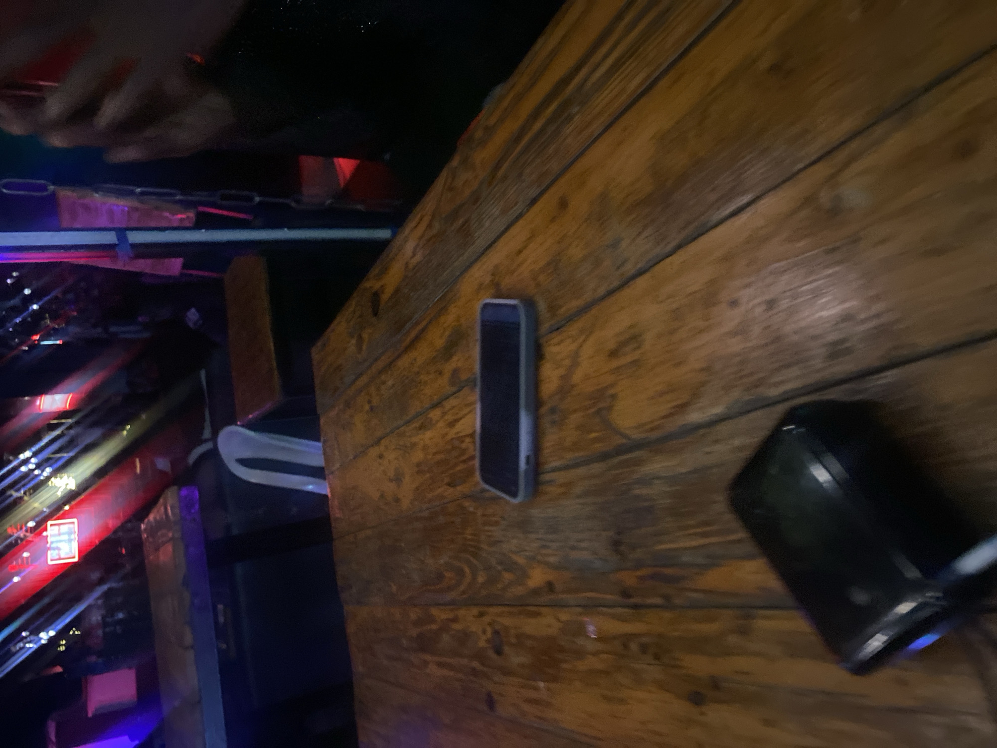 This image, captured in what is specified as Howayi, Nigeria, depicts an indoor, dimly lit recreational or social venue, likely a bar or club, during the evening or night. The foreground is dominated by a rustic wooden table constructed from multiple planks, exhibiting a warm brown tone with visible grain and minor wear. Centered on this table is a dark-colored smartphone with a light grey or light blue protective case, lying horizontally with its screen off. In the lower-left foreground, another dark, rectangular electronic device, possibly a power bank or a small communication device, is visible, displaying a faint blue light on its left side.
In the midground, beyond the primary table, additional dark-colored tables and chairs are discernable. A white plastic chair is positioned facing away from the viewer on the left side, behind another wooden table. The background is characterized by active artificial lighting, typical of entertainment venues. Beams of red, blue, and yellow-white light project from the upper-left, illuminating the dark space. A prominent rectangular red illuminated sign, possibly displaying text or a logo, is visible in the upper-center. To the far right, a vertical structure, potentially a pillar or wall segment, is adorned with hanging metal chains. Partially visible on the extreme right are a person's hands, seemingly holding a red object or illuminated by red light, alongside a dark, possibly textured sleeve, indicating the presence of an individual adjacent to the depicted scene. The overall environment suggests a vibrant, albeit underexposed, social setting.