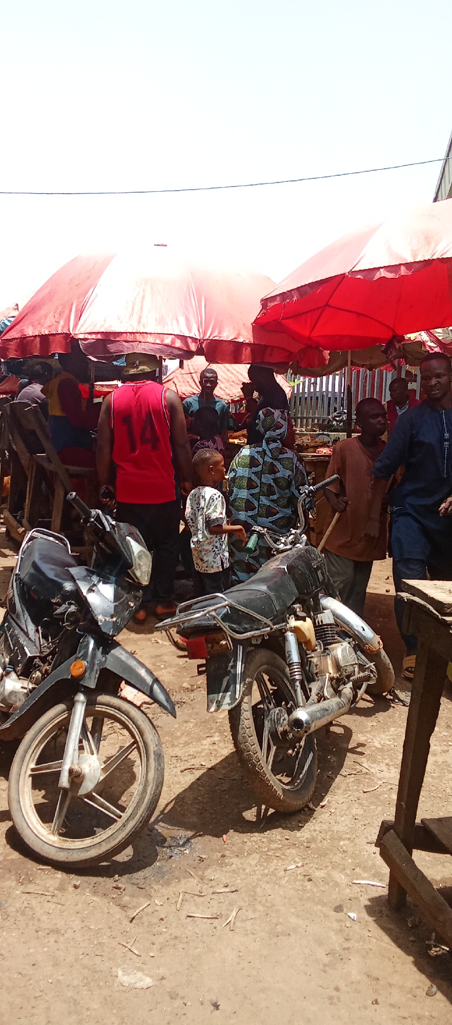 The image shows a bustling scene in Akwanga, Nigeria. Several individuals are gathered under large red umbrellas, suggesting a market setting. In the foreground, two motorcycles are parked on a dirt ground. A child in a light-colored patterned shirt stands among the crowd near the motorcycles. The surrounding area appears to be a market area with wooden tables and stalls. Several people are standing around. One man wears a red jersey with the number 14 on the back. There is a blend of casual clothing among the individuals, giving an impression of a typical day in a local market.