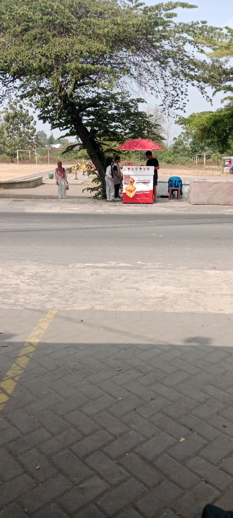 A street scene in Maguwoharjo, Indonesia, features a food cart with a red umbrella providing shade. A male vendor stands behind the cart, interacting with two female customers. A third woman stands to the left, observing the scene. In the background, a large tree with dense foliage dominates the upper left of the frame, with a children's playground visible further back. The foreground shows a paved road and sidewalk. The weather appears to be sunny.