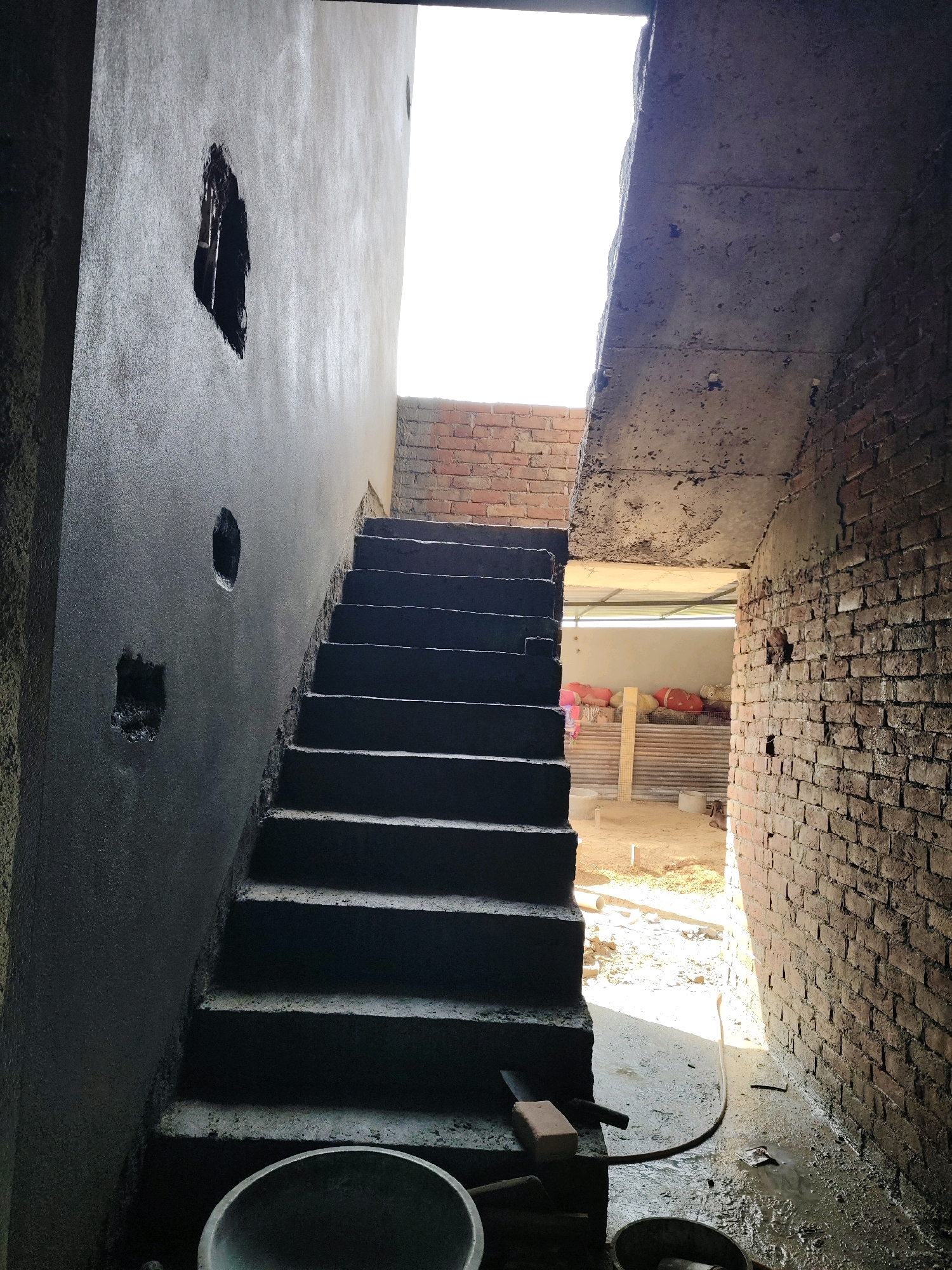 The image depicts an unfinished concrete staircase leading upwards, with a dark, possibly wet, wall on the left and a brick wall on the right. Several circular and rectangular openings are visible in the left wall. The area appears to be under construction, with exposed brickwork, concrete surfaces, and various construction materials scattered around, including bags of material, tools, and buckets. A hose lies on the wet ground near the base of the stairs. Sunlight streams in from an opening at the top of the stairs, illuminating the scene and casting shadows. The overall impression is one of an active construction site.