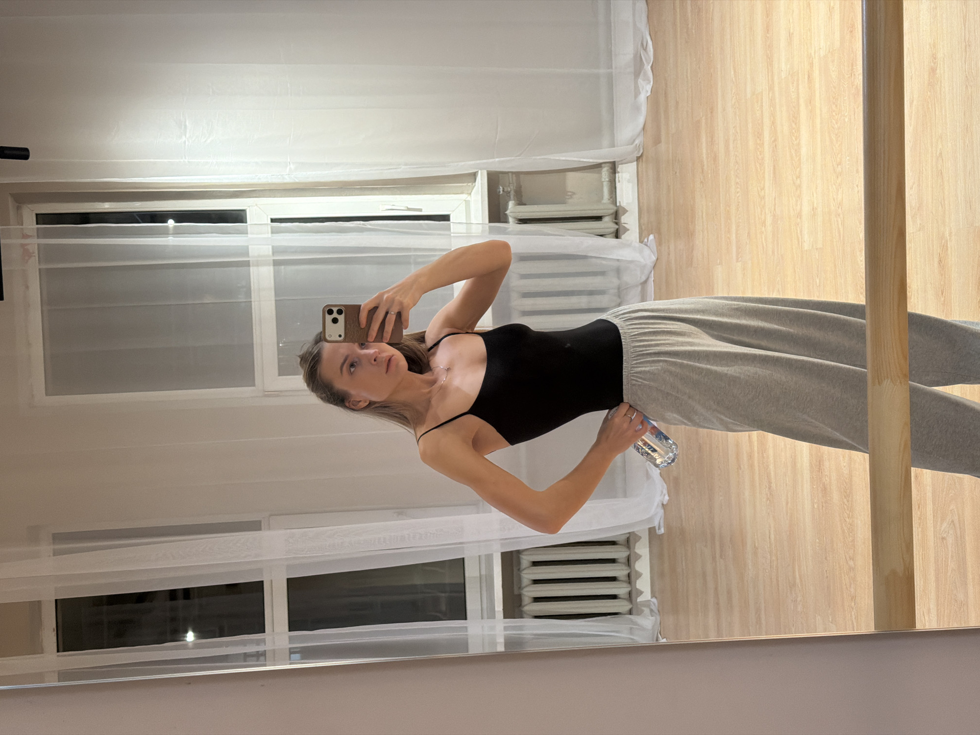 A woman is taking a selfie in a room. She is wearing a black top and gray sweatpants, and she is holding a water bottle. The room has two windows with white curtains, a radiator under each window, and a wooden floor. The scene appears to be in an indoor environment, potentially in a residential area. Moscow, Russia is not identifiable in the image.