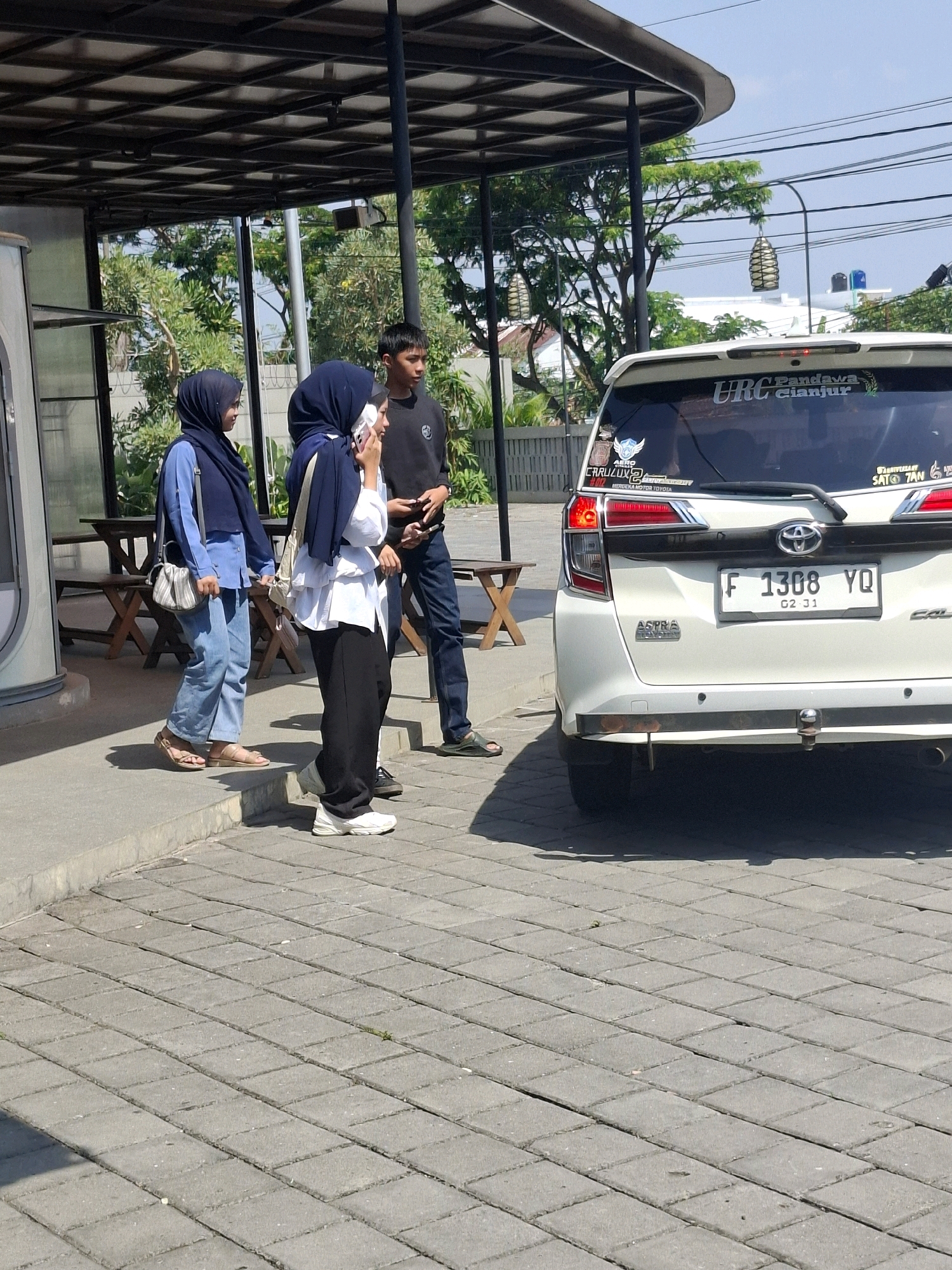 The image shows a street scene in Cianjur, Indonesia, on a sunny day. In the foreground, a white car is parked. The car has a license plate that reads "F 1308 YQ". Several stickers are visible on the rear window of the car, including one that says "URC Cianjur". Three people are standing on the sidewalk near the car. Two women, both wearing hijabs, are on the left. One woman is wearing a blue shirt and jeans, and the other is wearing a white shirt and black pants. A young man in a black shirt and blue jeans is standing to the right of the women, holding a mobile phone. In the background, there is a building with a covered outdoor seating area, trees, and power lines. The ground is paved with interlocking concrete blocks.
