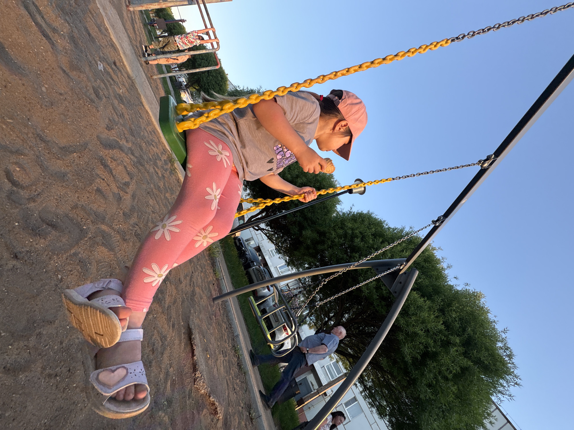 A young girl on a playground in Chaikovsky, Russia, is eating an ice cream cone while on a swing. She is wearing a pink baseball cap, a gray t-shirt, pink leggings with white daisy patterns, and sandals with heart embellishments. The swing has a green seat and yellow chains. The playground appears to be located near an apartment building with trees nearby. An adult male stands to the side, partially obscured by the swing structure. The time of day appears to be evening, with a clear blue sky.