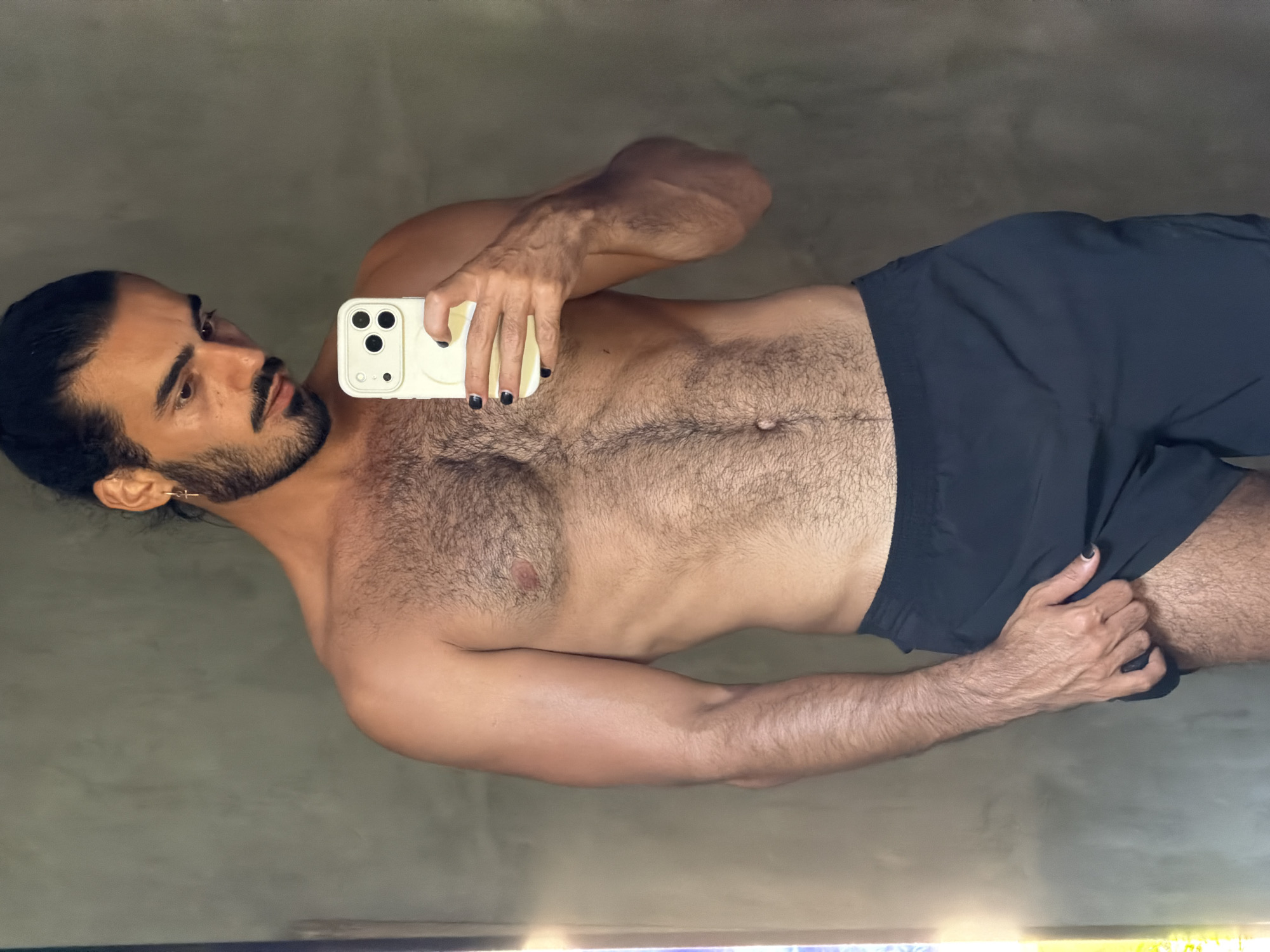 A shirtless man with dark hair pulled back in a bun, a beard, and a mustache, stands in front of a textured gray wall. He is wearing dark shorts and holding a light yellow smartphone in his right hand, taking a selfie. A thin gold cross earring is visible in his left ear. His chest and abdomen are covered in dark hair. His left hand is pulling up the hem of his shorts. A bright light source is visible to the left of the frame. The man appears to be indoors in a room with a concrete or stucco wall. The scene is devoid of any identifiable landmarks indicating a specific location in North Kuta, Indonesia, or any city.
