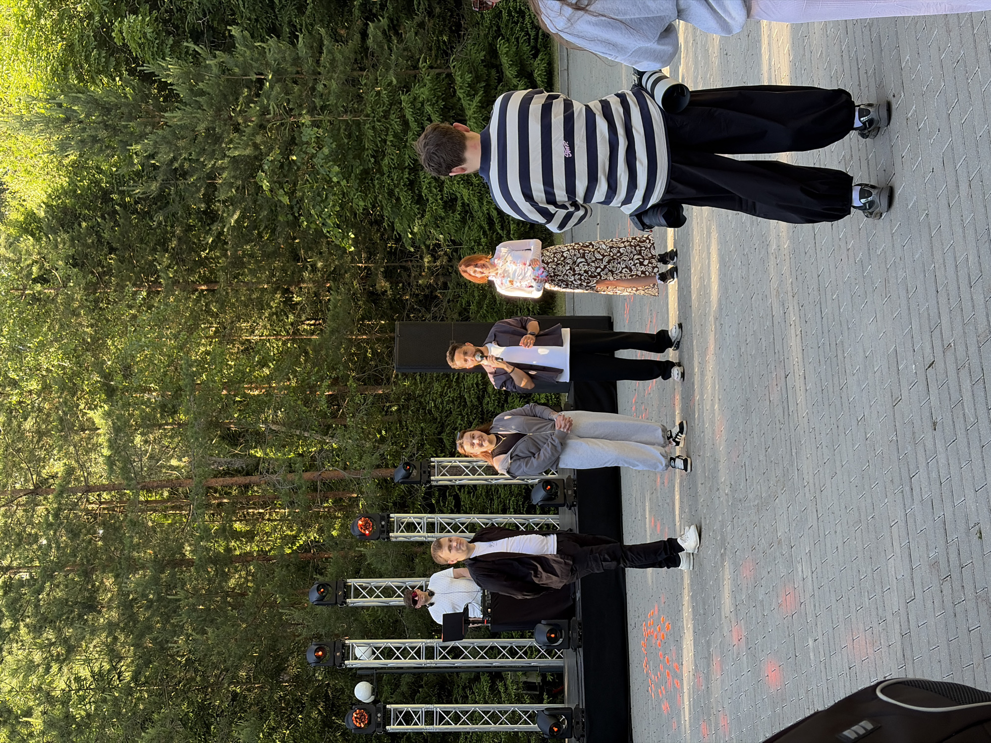 A group of people are gathered outdoors on a paved area in front of a line of trees. The sun is shining, casting shadows. Some people are standing in a line, facing towards the right of the frame. One person, wearing a black and white striped shirt and black pants, is facing away from the camera. Another person, in a white top and a patterned skirt, is smiling. Further down the line, a woman in a grey hooded jacket and light grey sweatpants is also smiling. At the front of the line, a man in a white shirt and black jacket is holding a microphone and appears to be speaking. Another man wearing sunglasses, a white shirt and dark pants, is standing next to him, appearing to be operating some equipment. Behind them, a stage structure with lights is visible. The overall scene suggests an outdoor performance or event. There is some visible text on the striped shirt. The location is Ждановіцкі сельскі Савет, Belarus.