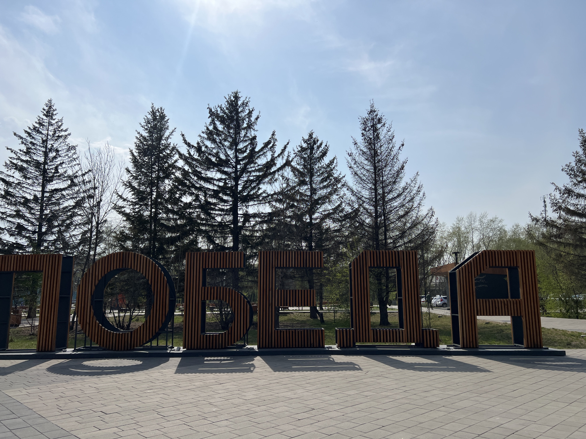 A wide-angle shot captures a large, modern wooden sign displaying the word "ПОБЕДА" (Victory in Russian), set against a backdrop of tall pine trees and a pale blue sky in Unknown, Unknown. The sign is composed of thick, vertical wooden slats with black accents, standing on a dark base that casts a shadow on the paved ground in front. The ground is covered with light grey, rectangular paving stones. The pine trees are dark green and dense, contrasting with the lighter sky. A hint of other greenery and structures can be seen through the gaps in the wooden sign and behind the trees. The sky is bright and slightly overcast.