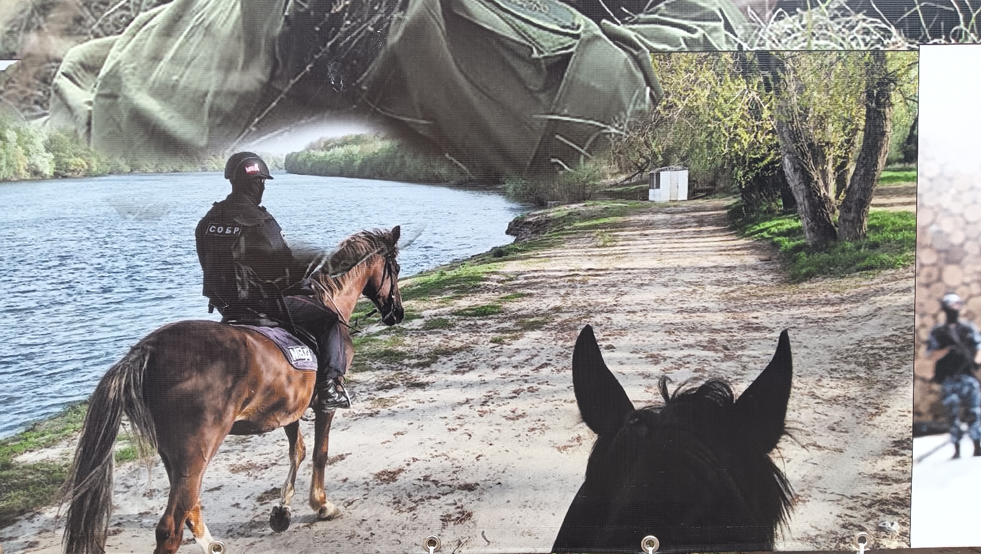 This image is a photograph of a large print or banner, indicated by visible grommets at the bottom, a seam-like vertical line on the right, and the printed texture. It depicts a tactical scene set in a natural environment, likely along a riverbank.
The central subject is a person in dark tactical gear, including a helmet, balaclava, and a protective vest, riding a brown horse. On the back of the vest, the Cyrillic acronym "СОБР" is clearly visible, signifying "Special Rapid Response Unit." The helmet and the horse's saddle pad also bear the Cyrillic acronym "МВД," which stands for "Ministry of Internal Affairs." The rider is positioned on the left side of a dirt or sandy path, looking towards a wide body of water, likely a river, that dominates the left half of the scene.
In the immediate foreground, the ears and head of a dark-colored horse are visible, suggesting the photograph is taken from the perspective of another rider. Along the right bank, there is green vegetation and trees with budding leaves. A small white structure, possibly a shed, is discernible in the distance among the trees. On the far right edge of the banner, a blurry, partially obscured figure in camouflage tactical gear is visible. The top portion of the banner features a textured, olive-green abstract pattern, resembling a folded piece of fabric or gear, rather than a natural sky.
The scene conveys an operational or surveillance activity during daytime. Given the location context of Tiraspol, Moldova, and the "СОБР" and "МВД" markings, the image points to a presence of Russian-affiliated or Transnistrian special police/military forces, possibly conducting a patrol along the Dniester River.
