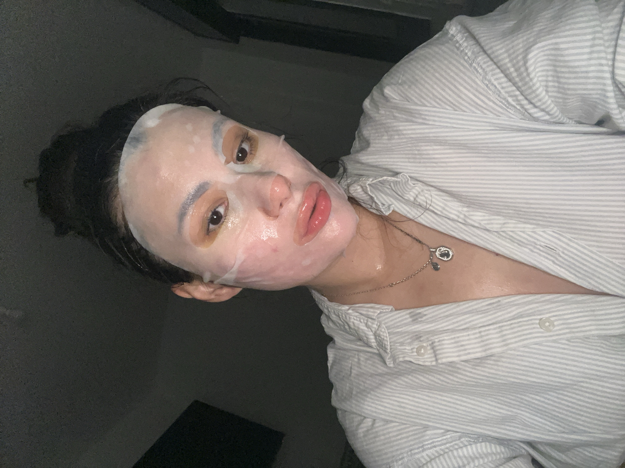 A single individual is depicted in a close-up, dimly lit indoor setting identified as Kiryat Bialik, Israel. The subject, an adult female, is facing the camera directly, capturing what appears to be a selfie. Her dark hair is pulled back, and her face is covered by a translucent sheet mask, which has cutouts for the eyes, nostrils, and mouth. The mask appears moist, with small surface irregularities. Her eyes are open, revealing dark irises and eyelids adorned with a brownish-gold eyeshadow. Her lips are visible through the mask's opening, appearing full and glossy with a reddish tint. The skin around the mask and visible areas shows a slightly flushed complexion. She is wearing a light-colored, vertically striped, collared button-down shirt, unbuttoned to reveal the upper chest. A silver-colored chain necklace with two pendants is worn around her neck; one pendant is small and dark, the other is larger, oval-shaped, and features a detailed design. The background is dark and largely undifferentiated, with a dark rectangular object partially visible on the left side.