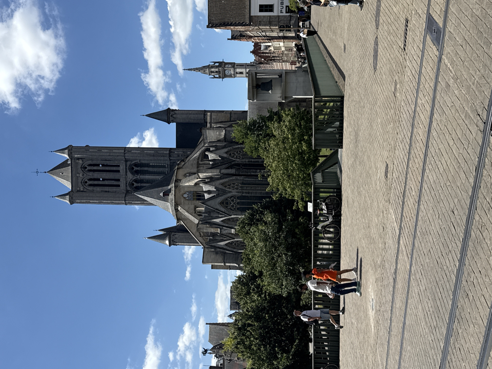 A vertical medium shot captures a scene in Ghent, Belgium, on a sunny day with scattered clouds. The focal point is the imposing Gothic architecture of a large church, likely St. Bavo's Cathedral, with its prominent towers and intricate stone carvings. Adjacent to the church, a tall, slender clock tower with visible clock faces stands out against the blue sky.
In the foreground, a paved walkway made of grey bricks runs diagonally across the frame. A group of four people are walking on this path. The first person from the right wears a white t-shirt and dark shorts. Next to them, a person in an orange top and dark pants walks ahead. Following them are two more individuals, one in a white t-shirt and jeans, and another in a grey t-shirt and shorts. They appear to be casually strolling.
To the left of the walkway, lush green trees partially obscure the view of the church. A green metal fence or railing separates the walkway from the area with the trees and church. A bicycle is visible leaning against the railing.
The lighting suggests it is daytime, likely mid-morning or early afternoon, given the clear blue sky and the shadows cast by the people. The overall atmosphere seems peaceful and pleasant, characteristic of a tourist-friendly location. No discernible text is visible in the image.