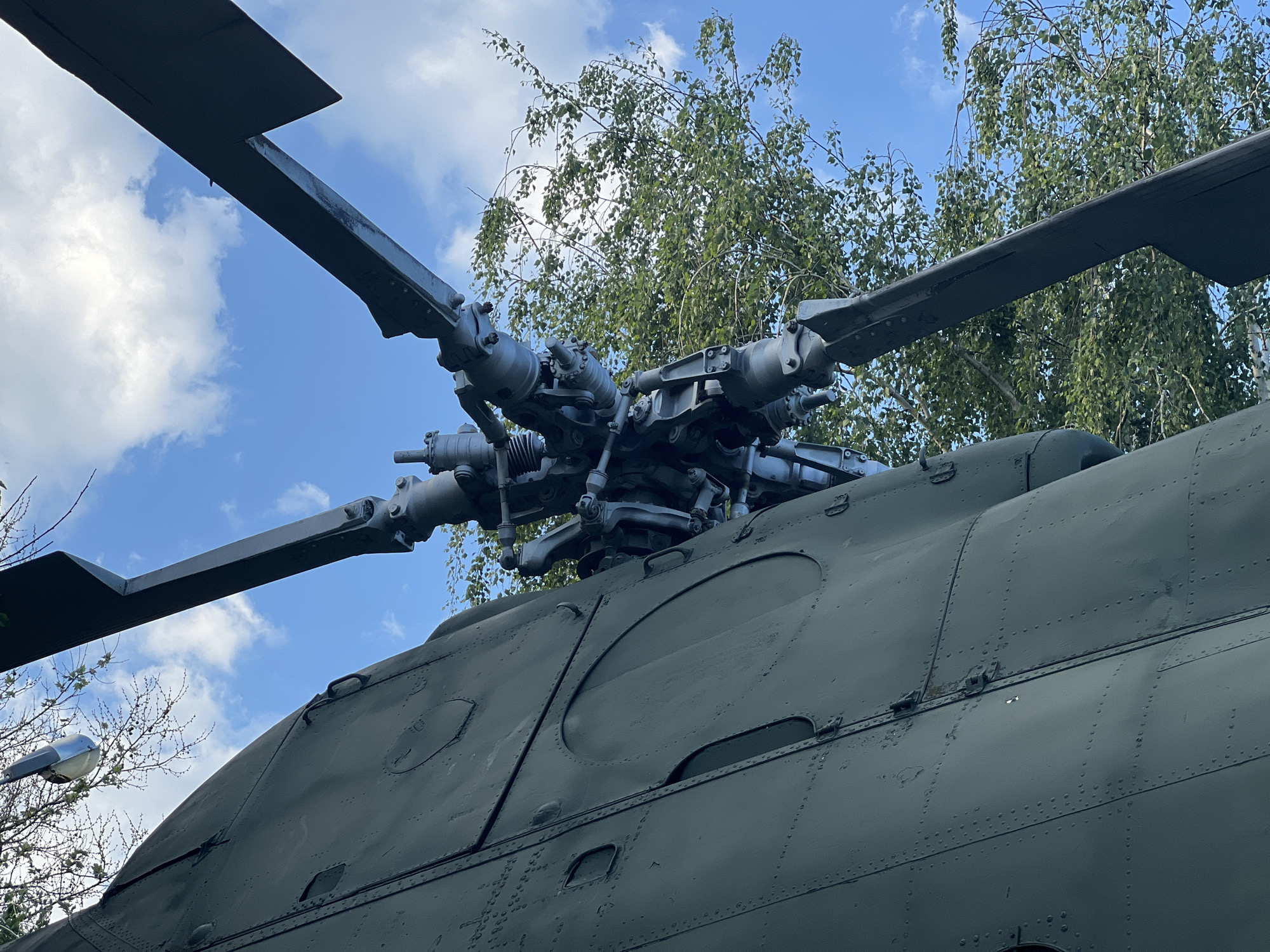 The image depicts the rotor and upper fuselage of a Mil Mi-8 helicopter. The helicopter is painted in a matte green color, exhibiting rivets and panel lines. The rotor blades are gray and positioned against a backdrop of a blue sky with scattered clouds and the green foliage of a tree. A street lamp is visible to the bottom left corner of the photo. This particular helicopter is located at the Park of the 30th Anniversary of the Victory in Krasnodar, Russia.